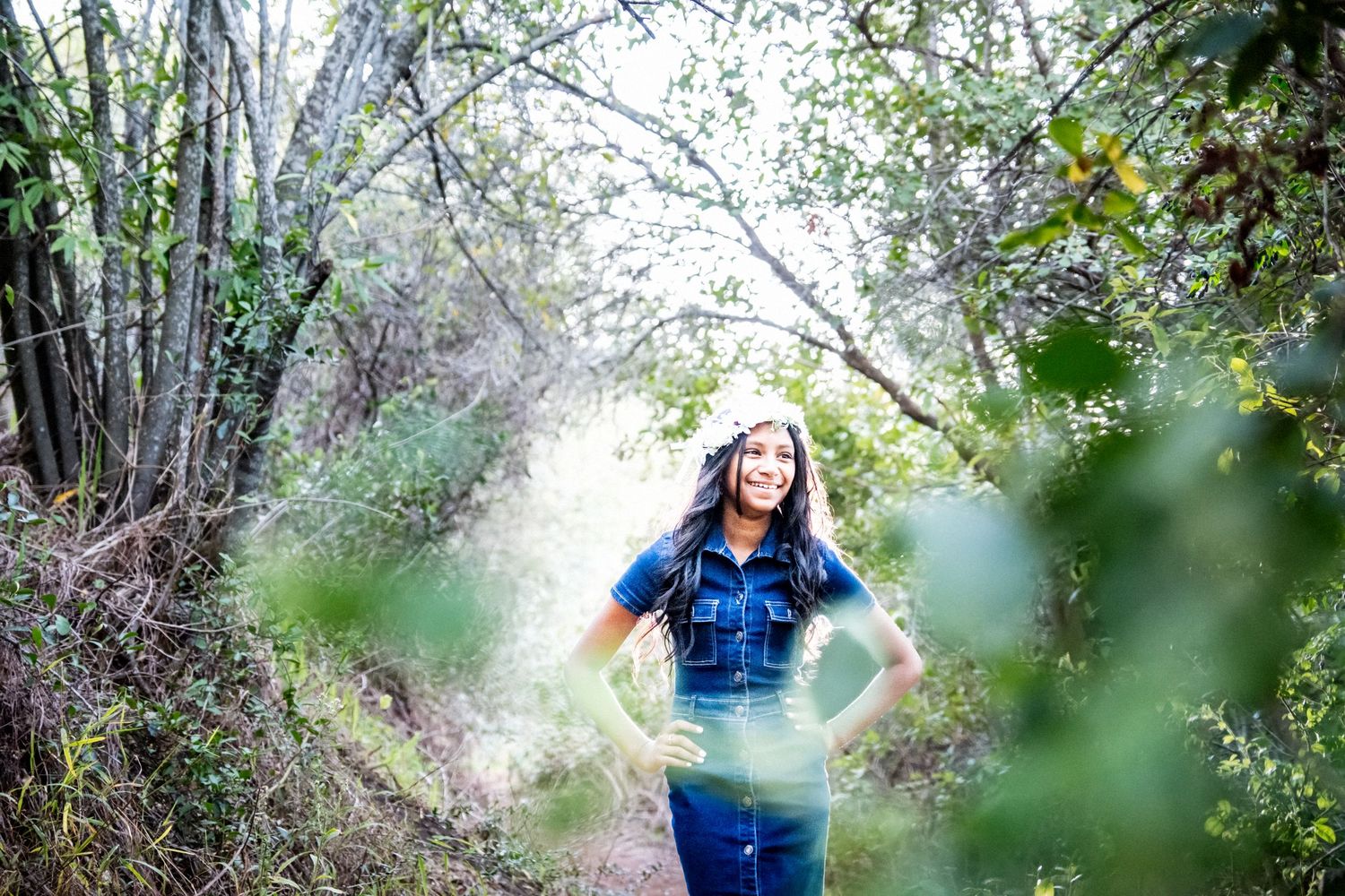 Woman in denim outfit smiling on forest trail surrounded by lush green foliage.