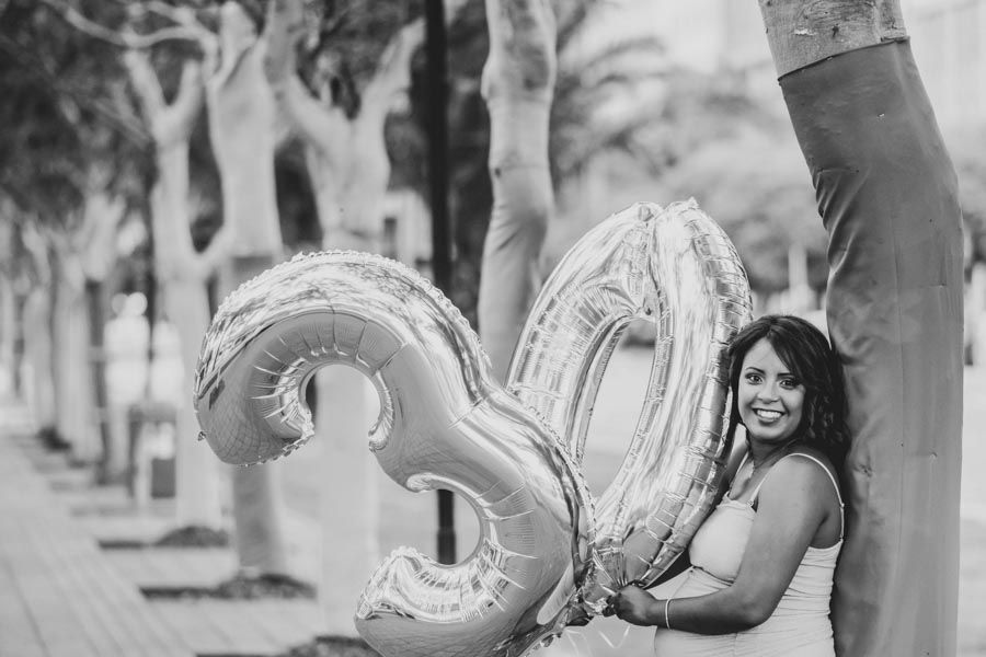 Black and white photo of smiling woman holding large number 30 balloons outdoors among trees.
