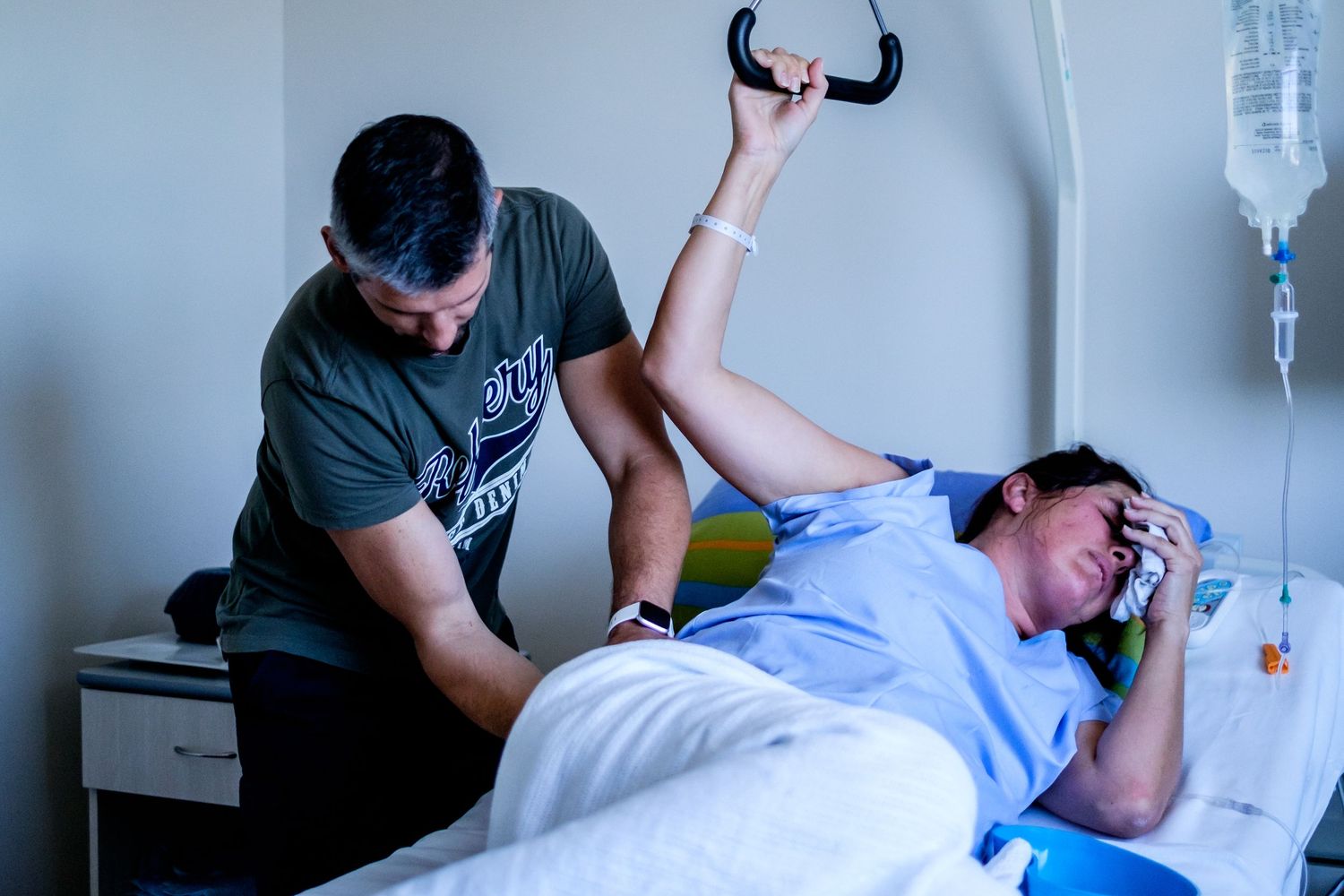 A woman in labor grips a handle in a hospital bed while a supportive partner assists her during childbirth.