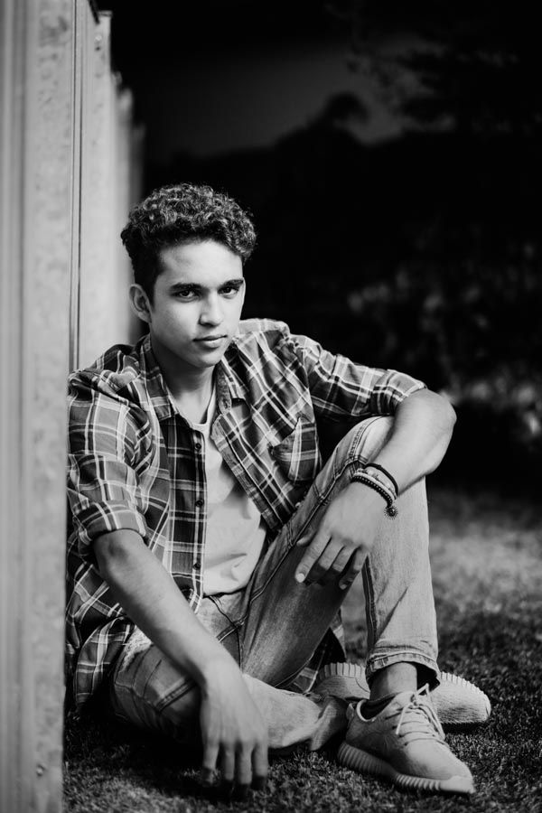 Black and white portrait of a young man in plaid shirt sitting against a wall outdoors for his model portfolio shoot.