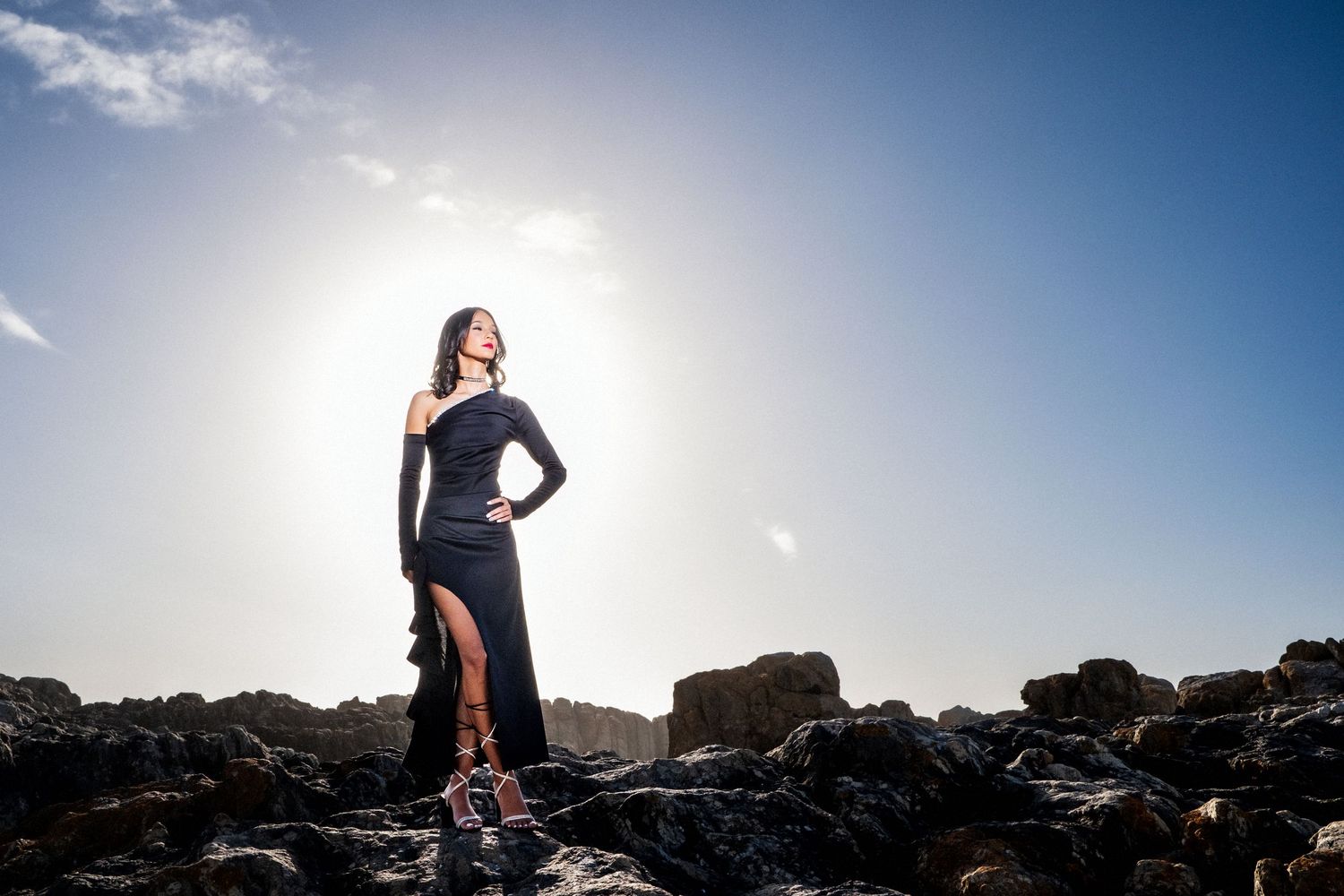 Woman in black dress posing dramatically on rocky coastline against bright sunny sky.