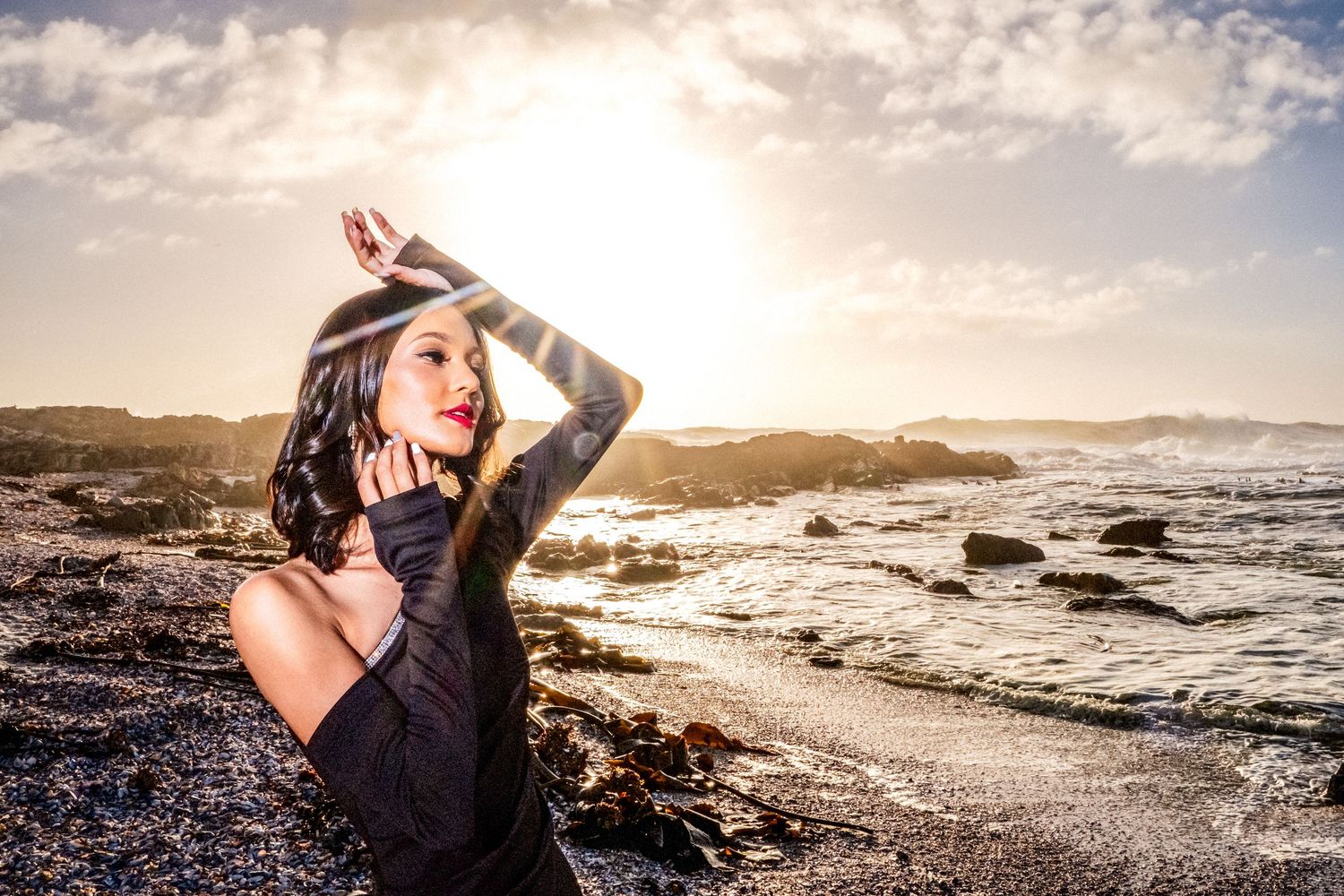 Woman in black dress posing on sandy beach with ocean waves and golden sunset light.