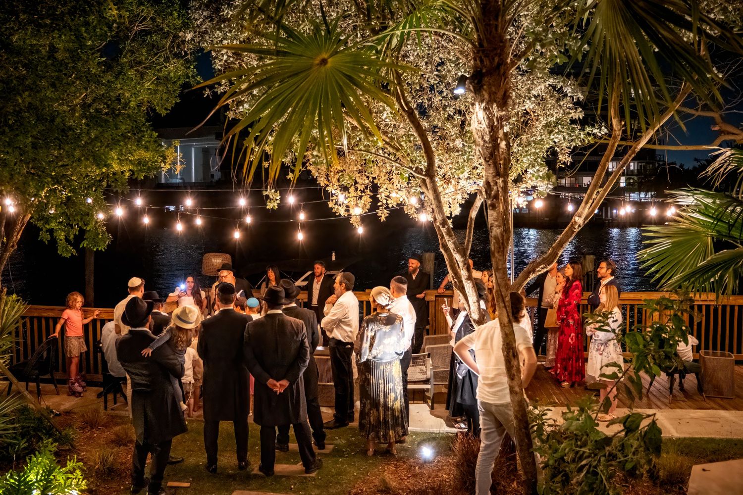 Guests mingling at an elegant outdoor evening party with string lights and tropical trees.