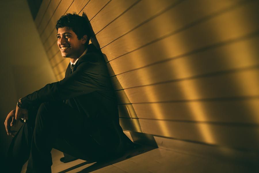 Young teenager male in dark suit sitting against a wall in dramatic golden shadow lighting for his senior portraits photoshoot.
