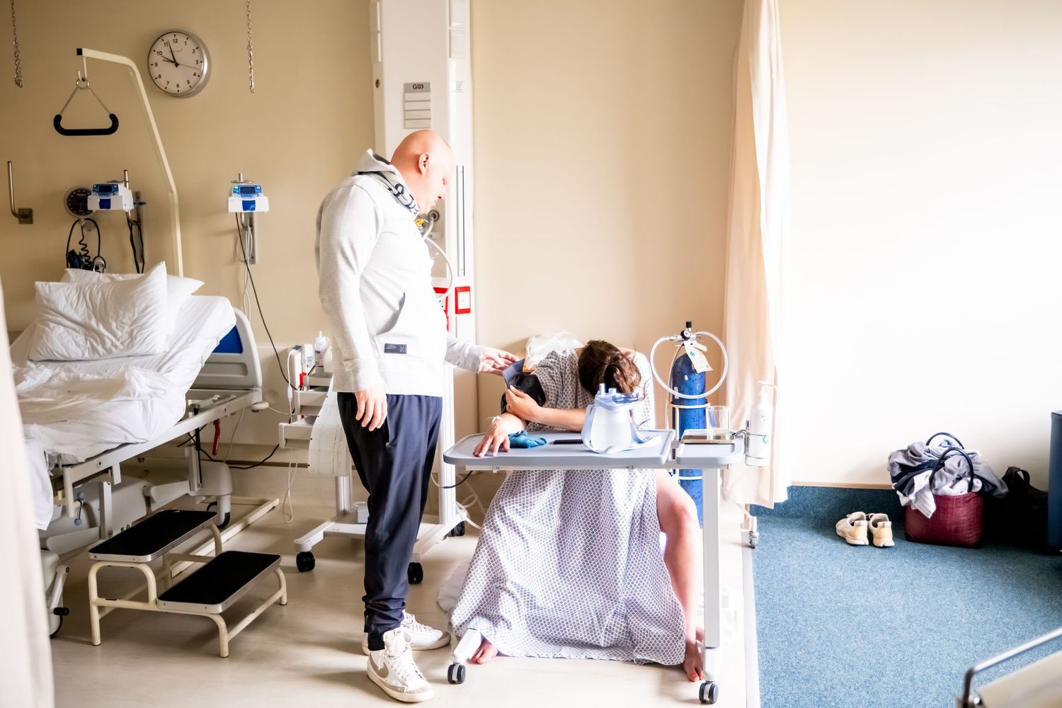 A patient in a hospital gown sits at a bedside table while a visitor stands nearby in a medical room.