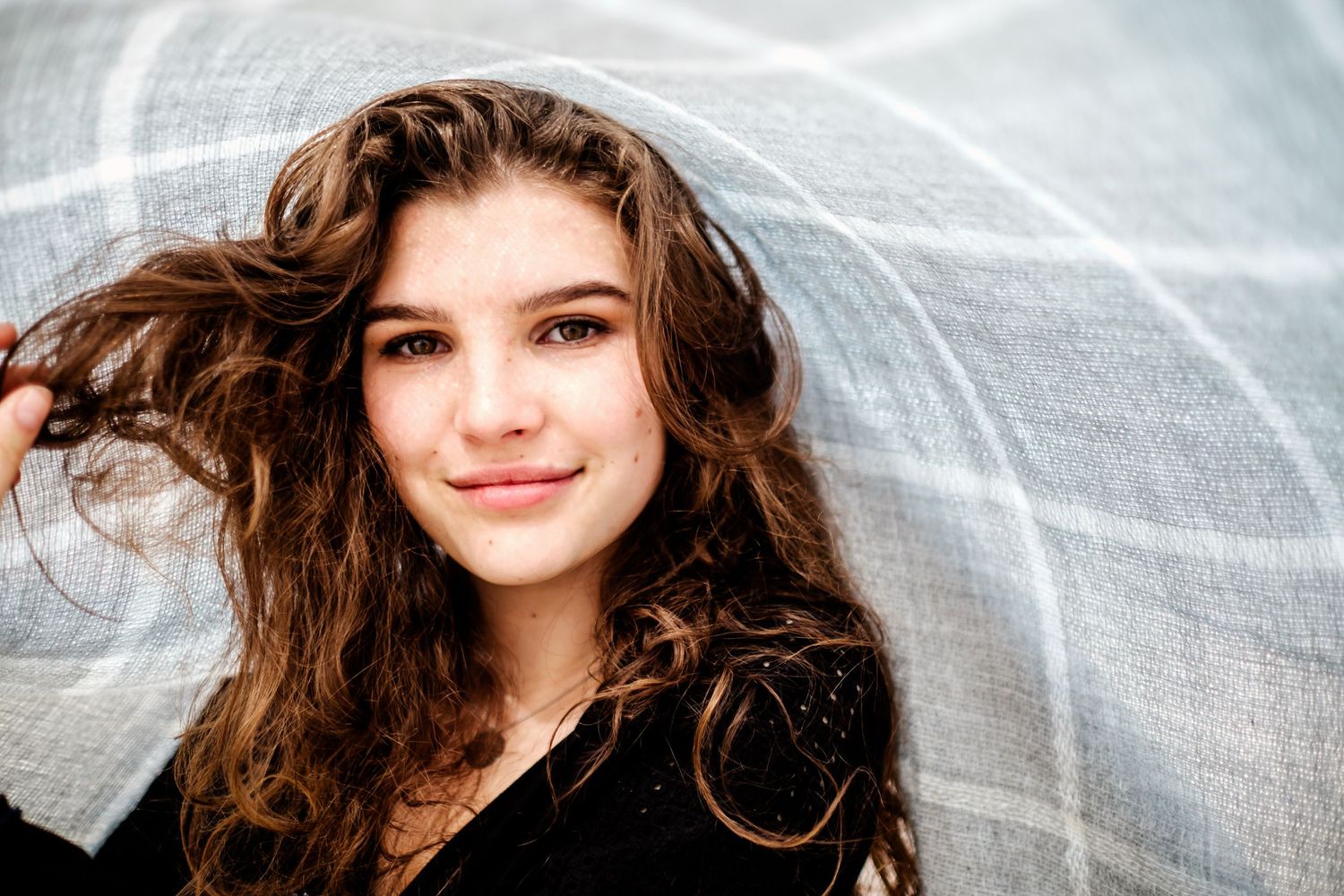 Smiling teenage girl with curly brown hair posing under a sheer white fabric outdoors for her teen model shoot.