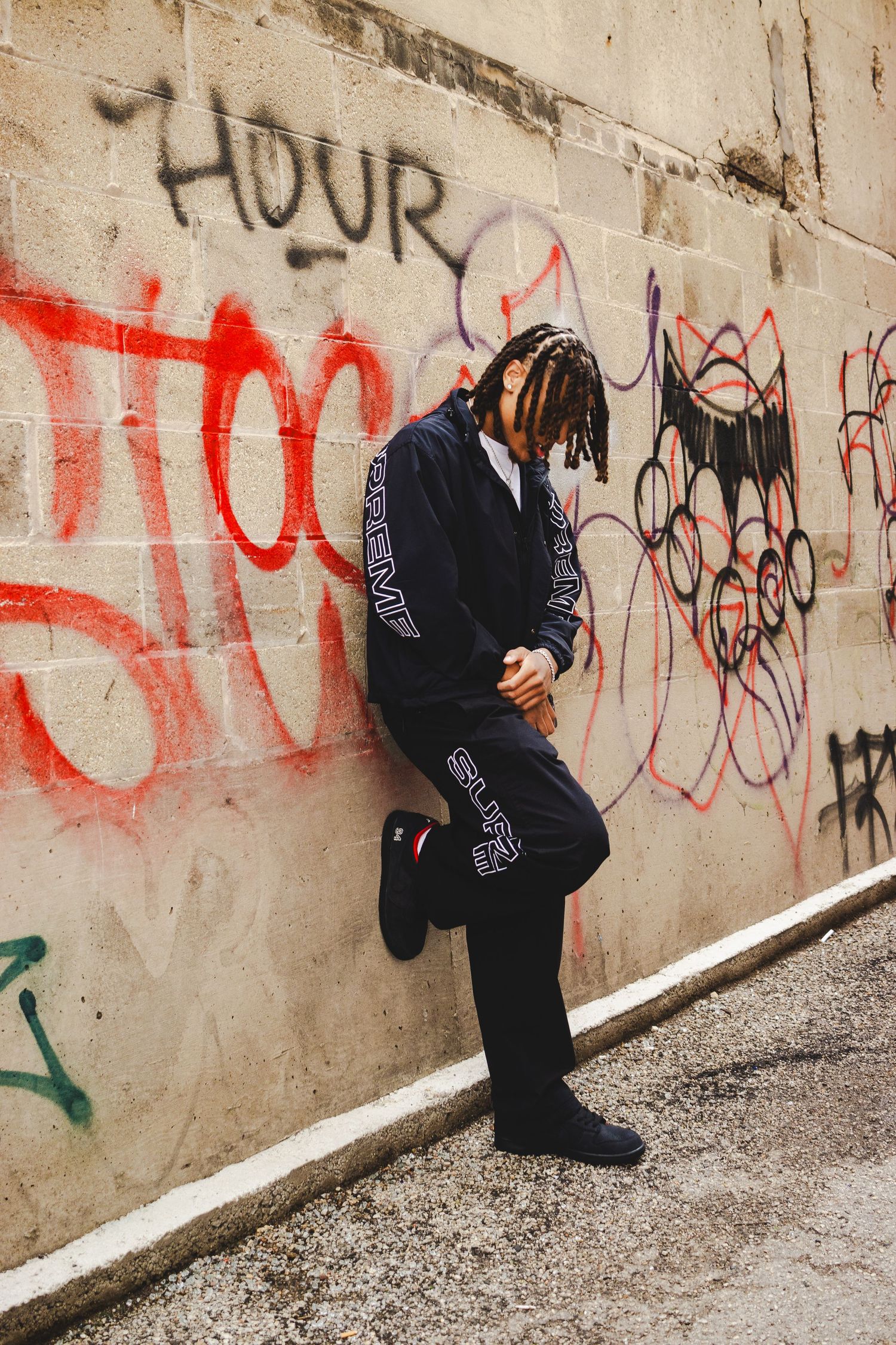 Person in dark clothing poses near industrial wall with street art.