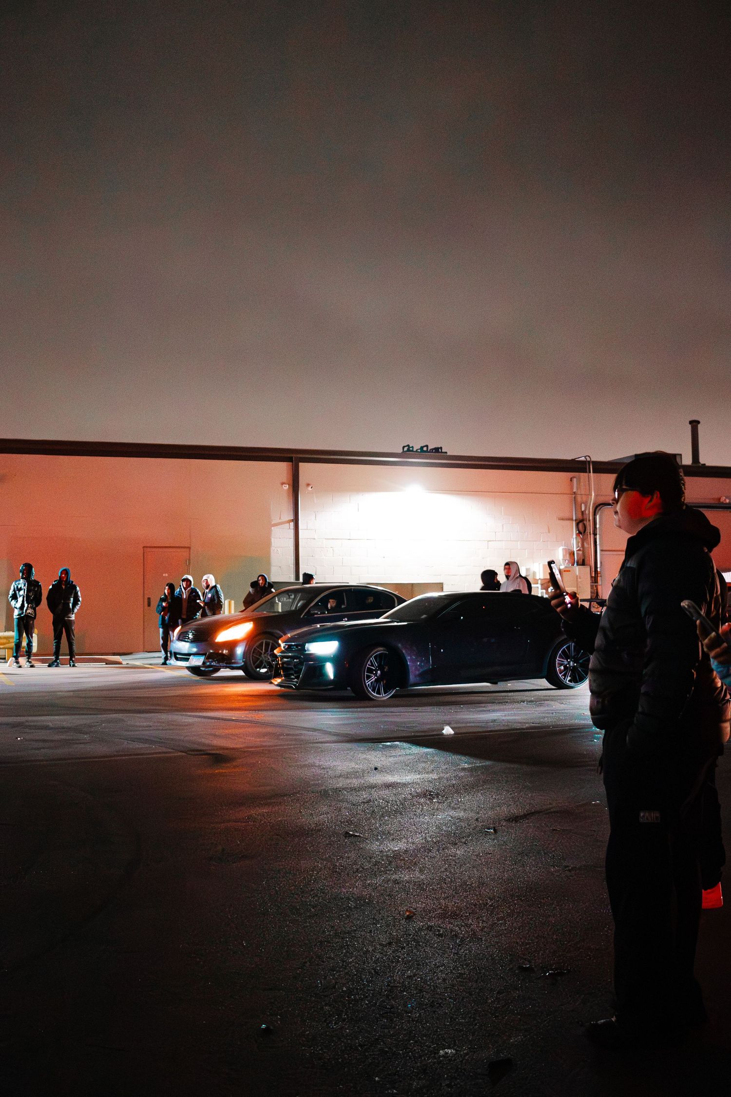 Nighttime gathering of cars and people in a dimly lit parking lot.