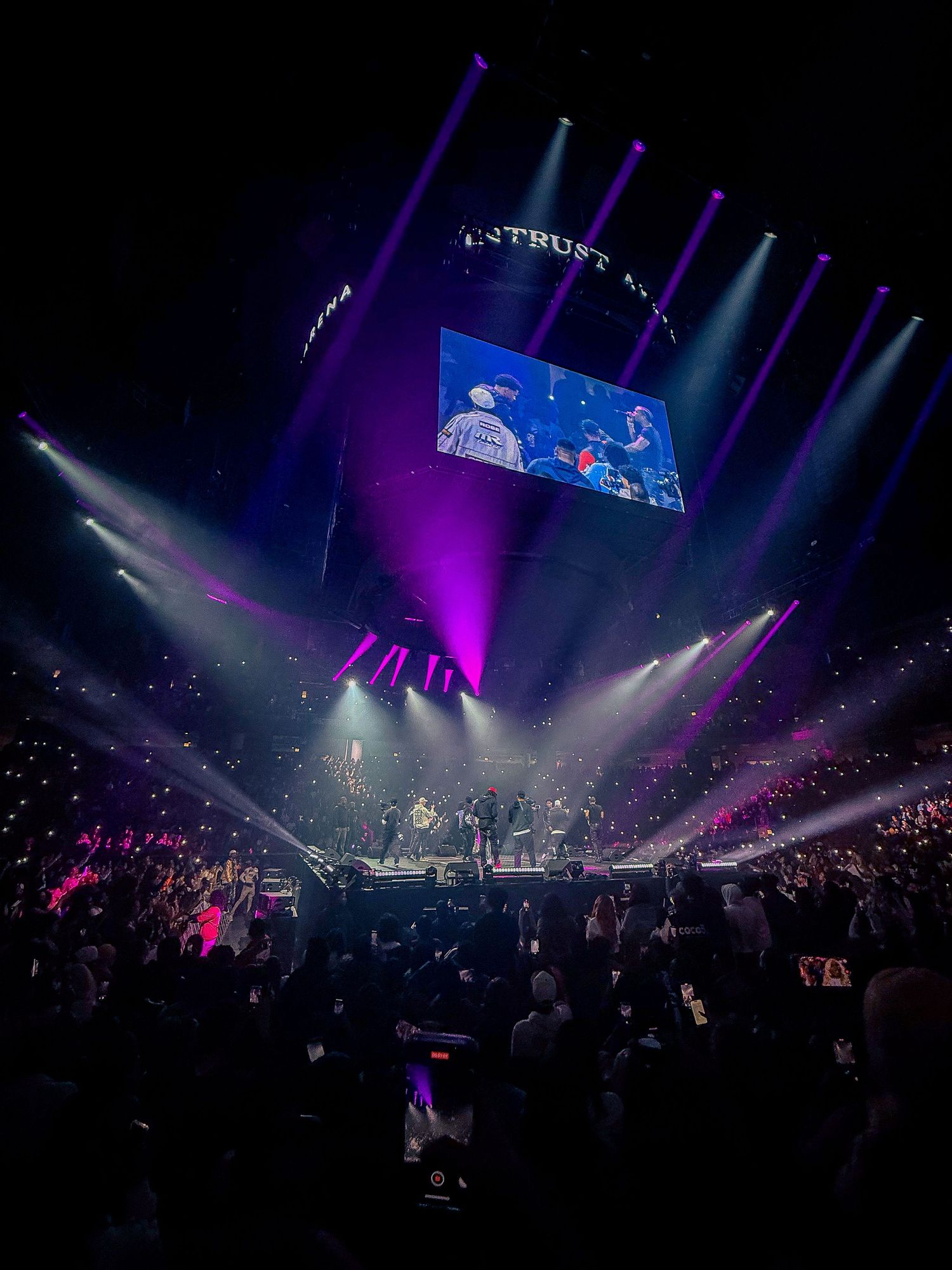 Concert arena illuminated with purple lights and fog effects showing performers on stage with large video screen display above.