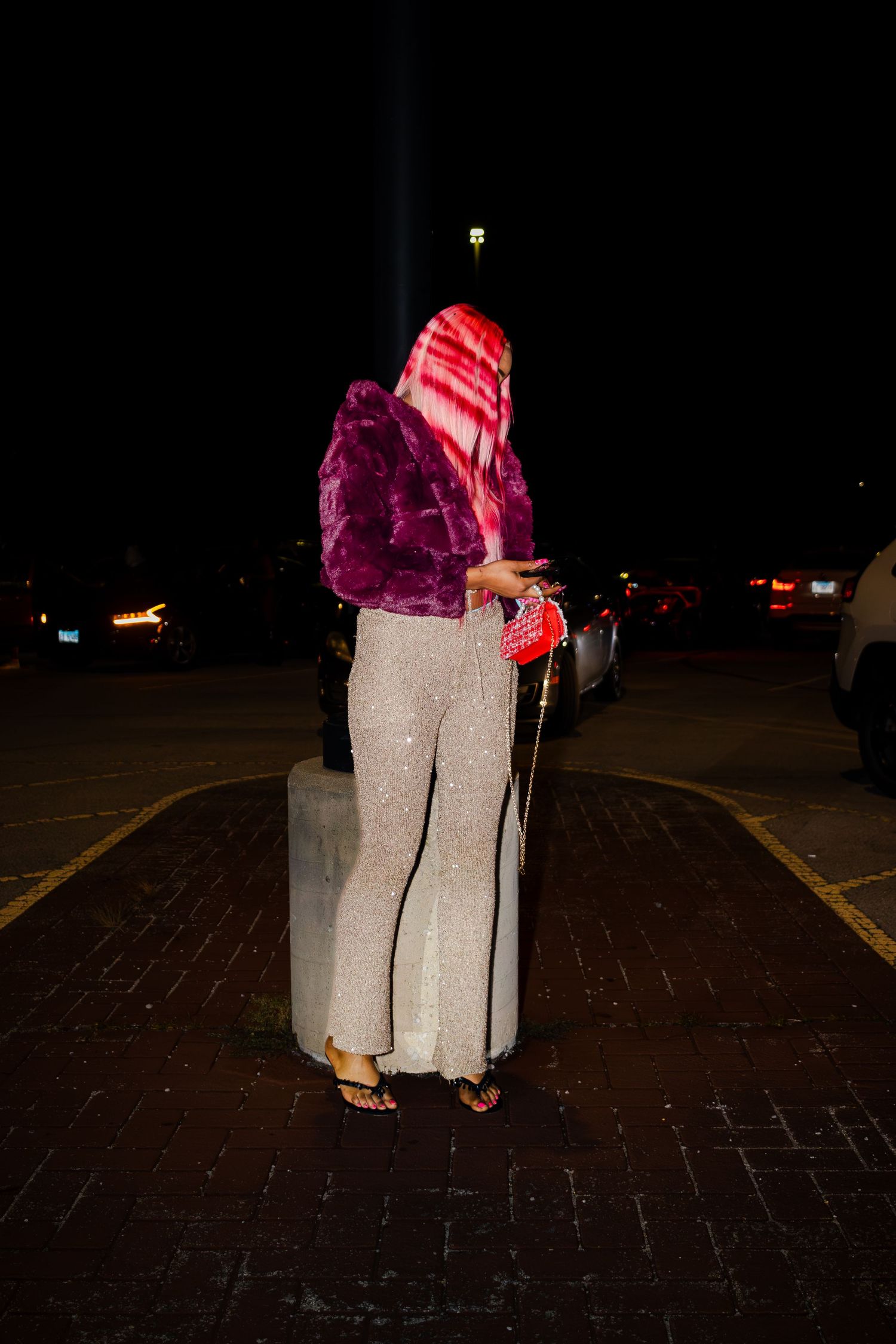 Figure in sparkly dress and pink accessories stands on brick pavement at night.