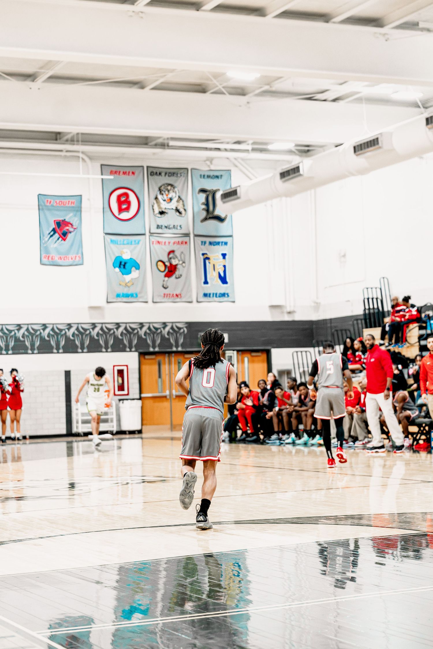 High school basketball game in progress with players on court and championship banners hanging on gymnasium wall.