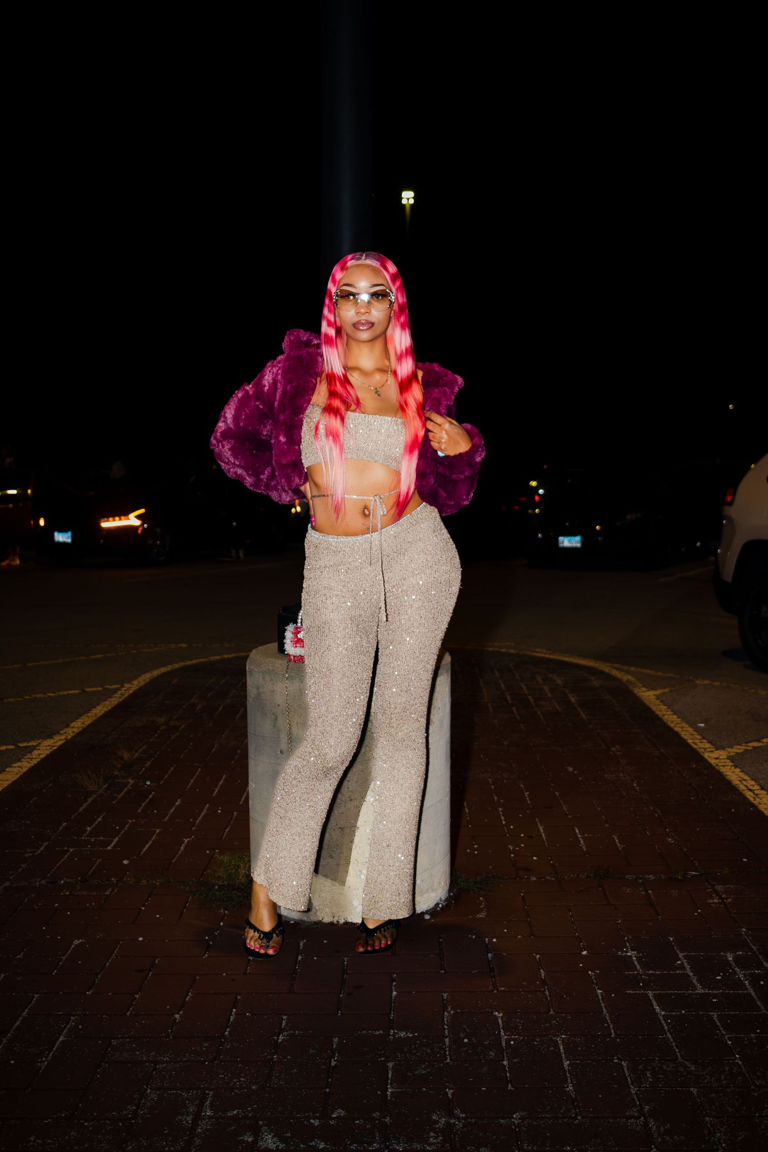 A person in a pink fur jacket and sparkly pants poses at night on a brick surface.