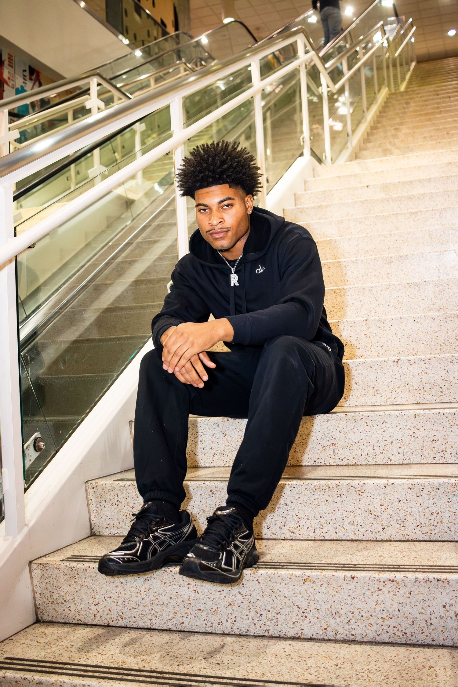 Someone in casual black athletic wear sits on marble stairs near an escalator in a modern indoor setting.