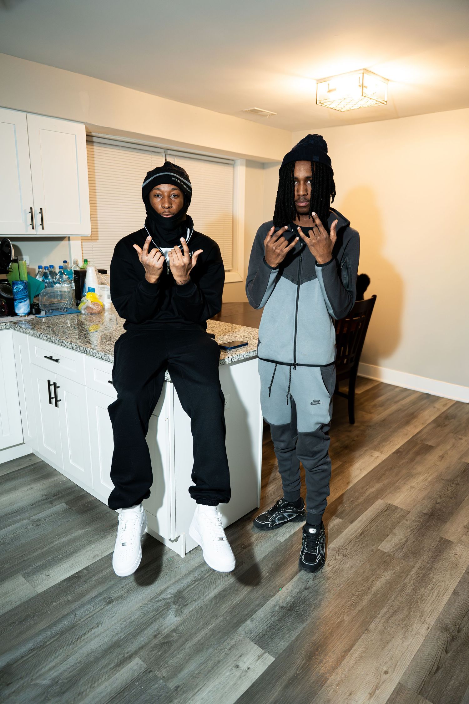 Two people in casual streetwear stand in front of white kitchen cabinets and hardwood floors.