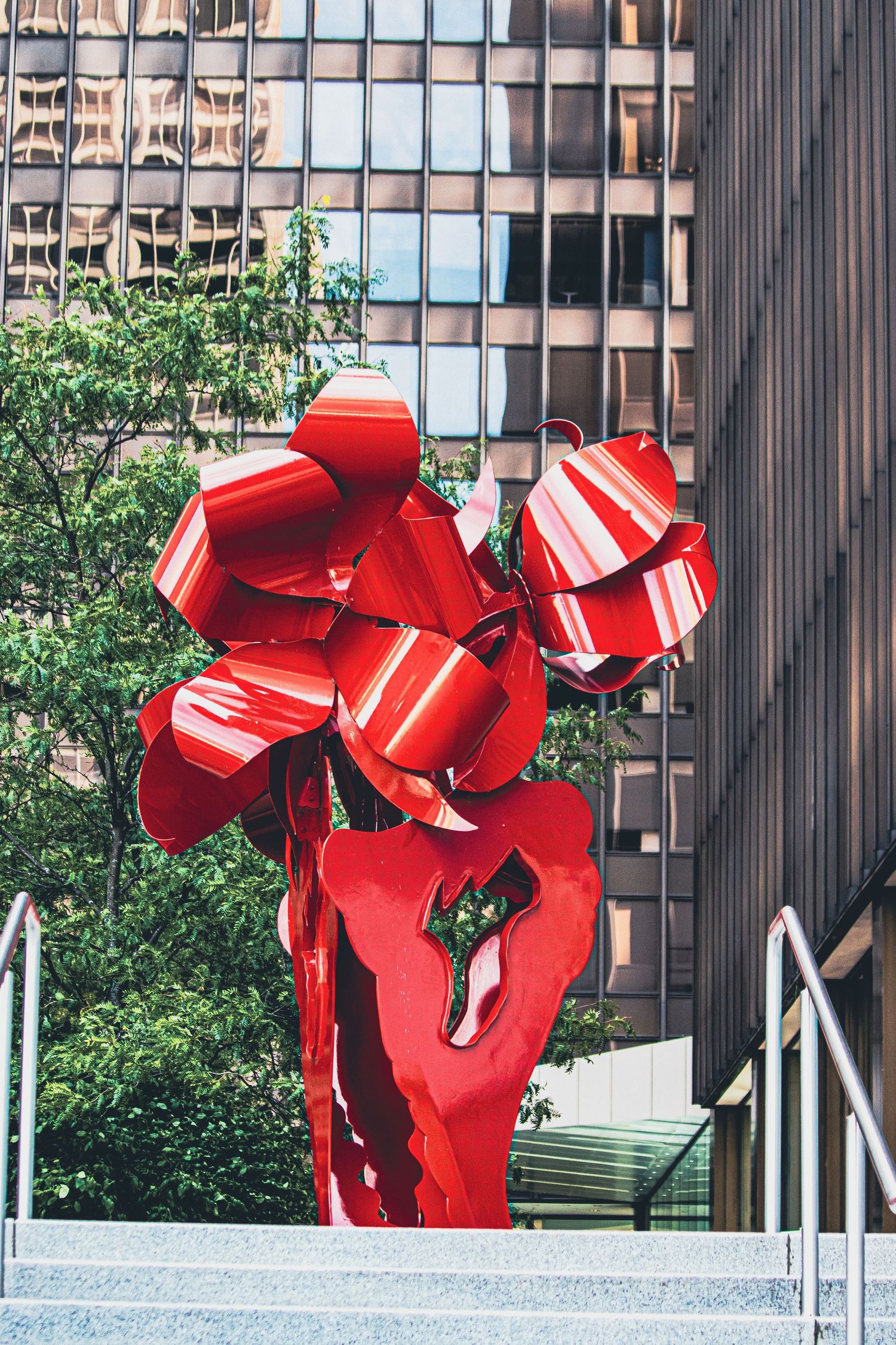 Large red abstract metal sculpture installation with angular geometric forms displayed in an urban setting with glass buildings.
