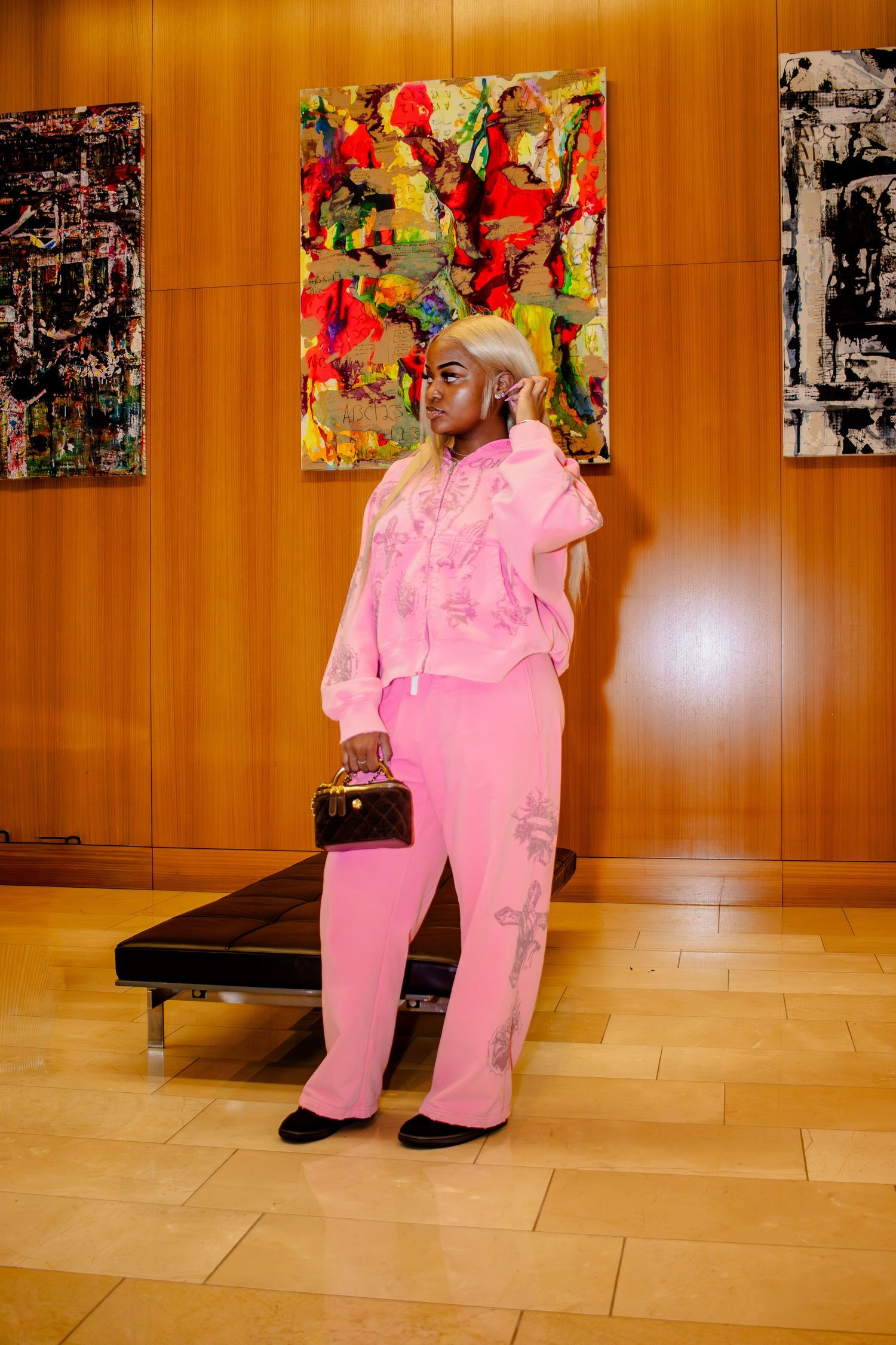 A person wearing a bright pink jumpsuit and red floral headpiece poses in an art gallery with paintings on wooden walls.