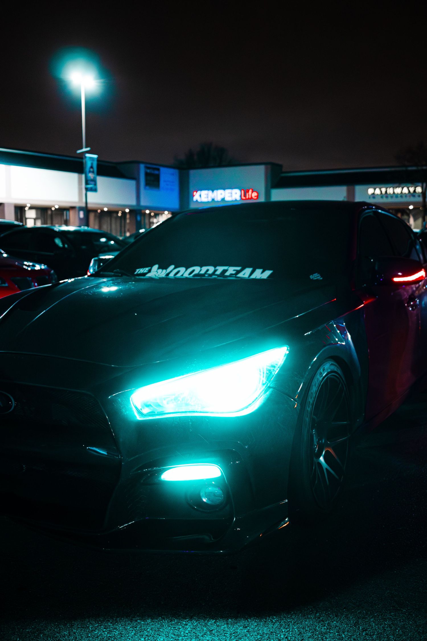 A sports car with glowing blue headlights and illuminated logo parked in a dark parking lot at night.