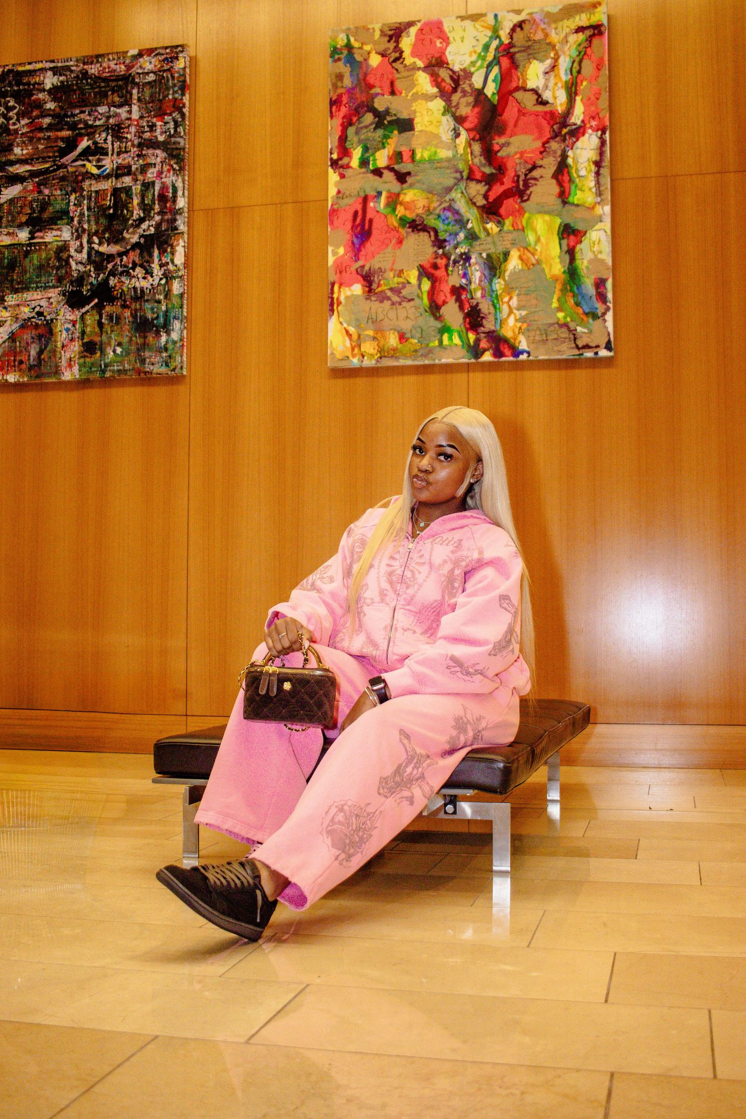 A figure in a pink robe sits on a wooden floor against warm wood paneling with colorful abstract artwork displayed above.