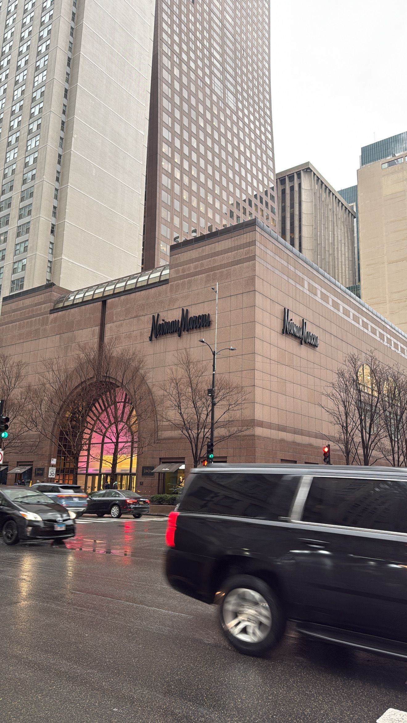 A series of black vehicles pass by a Neiman Marcus department store exterior on a rainy day.