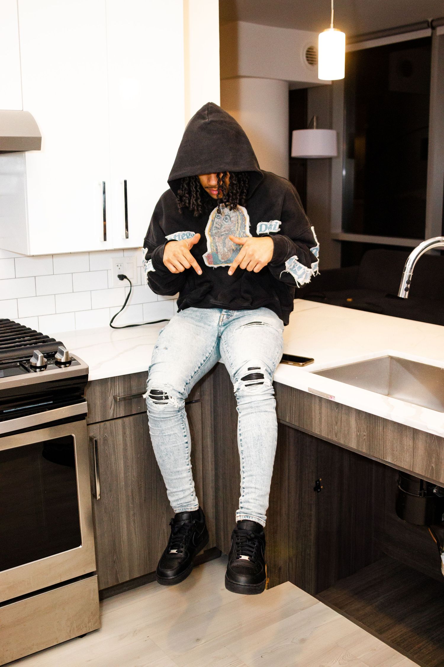 Person wearing distressed jeans and black hoodie sits on modern kitchen counter with white tile backsplash.