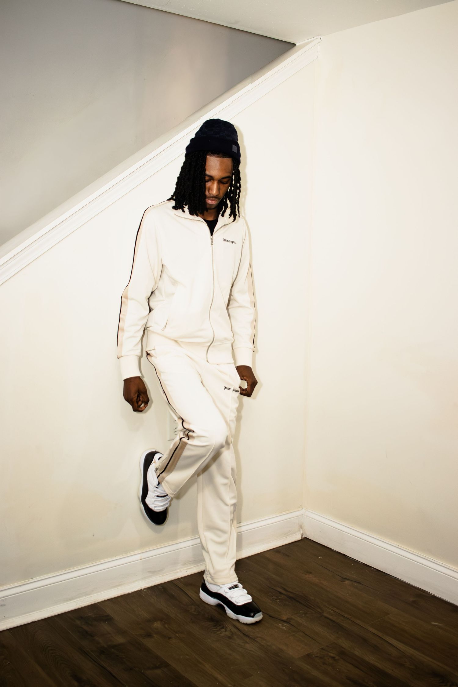 A person in a crisp white tracksuit and sneakers poses against a white wall on hardwood floors.