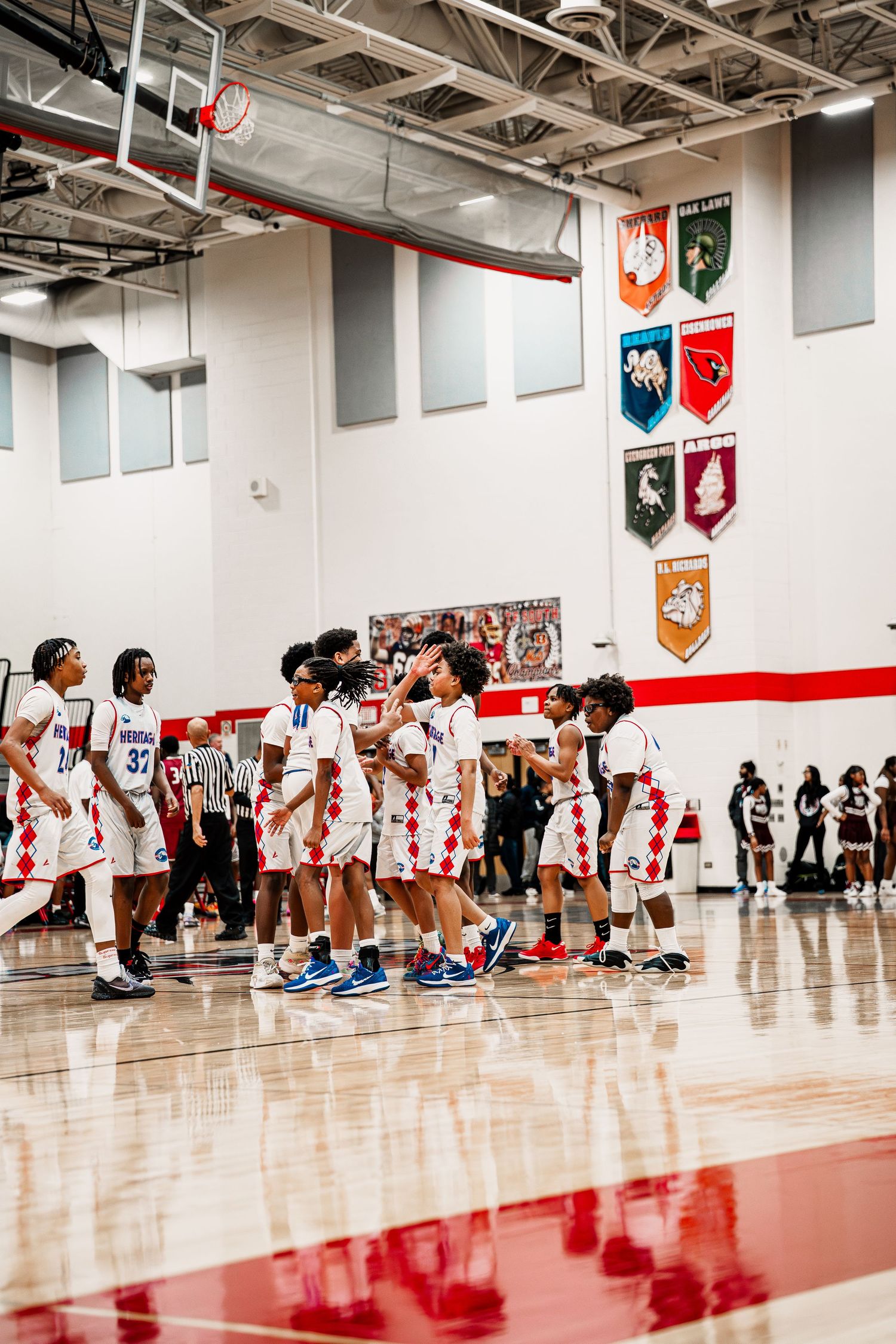 Students line up along a gym wall during a basketball practice session on a shiny red court.
