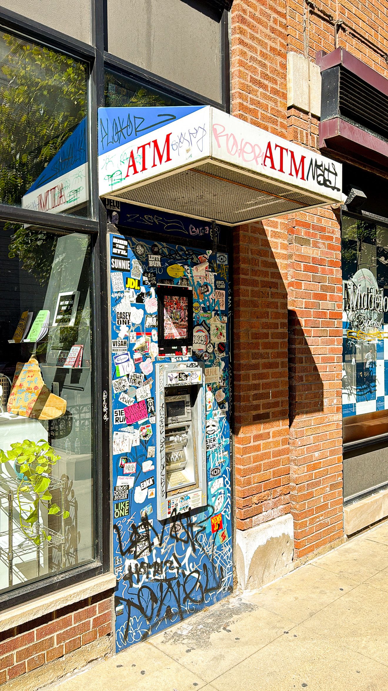 ATM machine on brick wall covered in graffiti and stickers outside urban storefront.