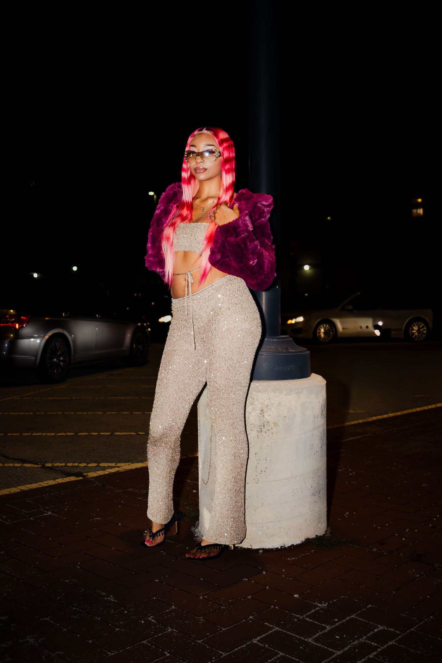 A figure in a sparkly silver dress and pink hair poses dramatically in a dark parking lot at night.