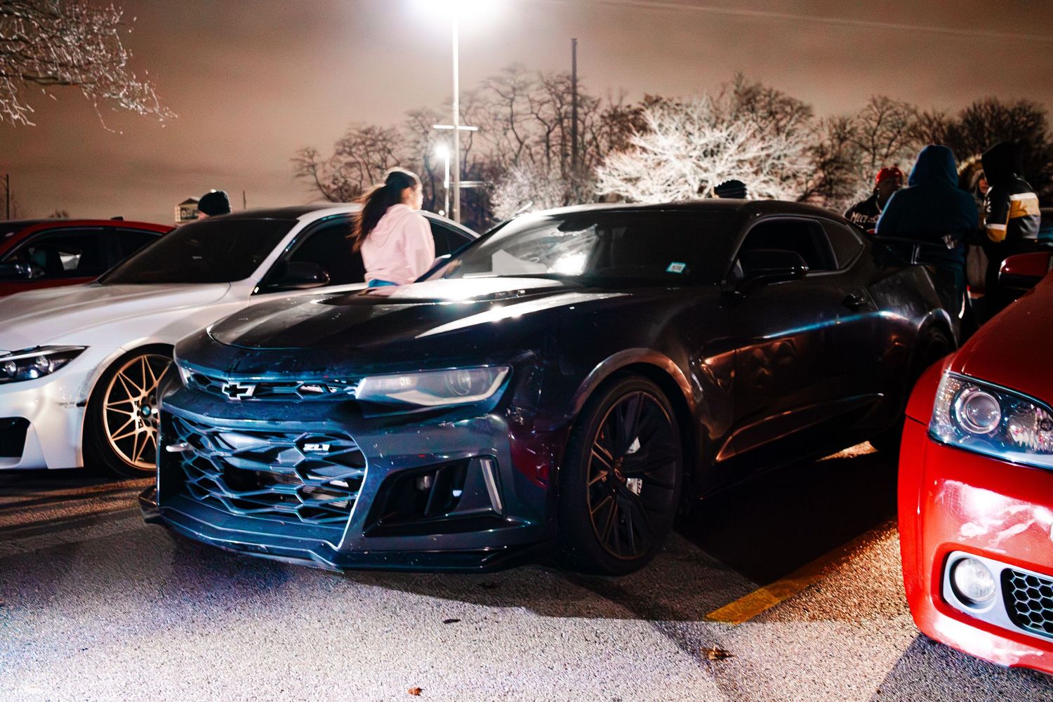A row of modern sports cars and luxury vehicles parked at night under bright street lighting.