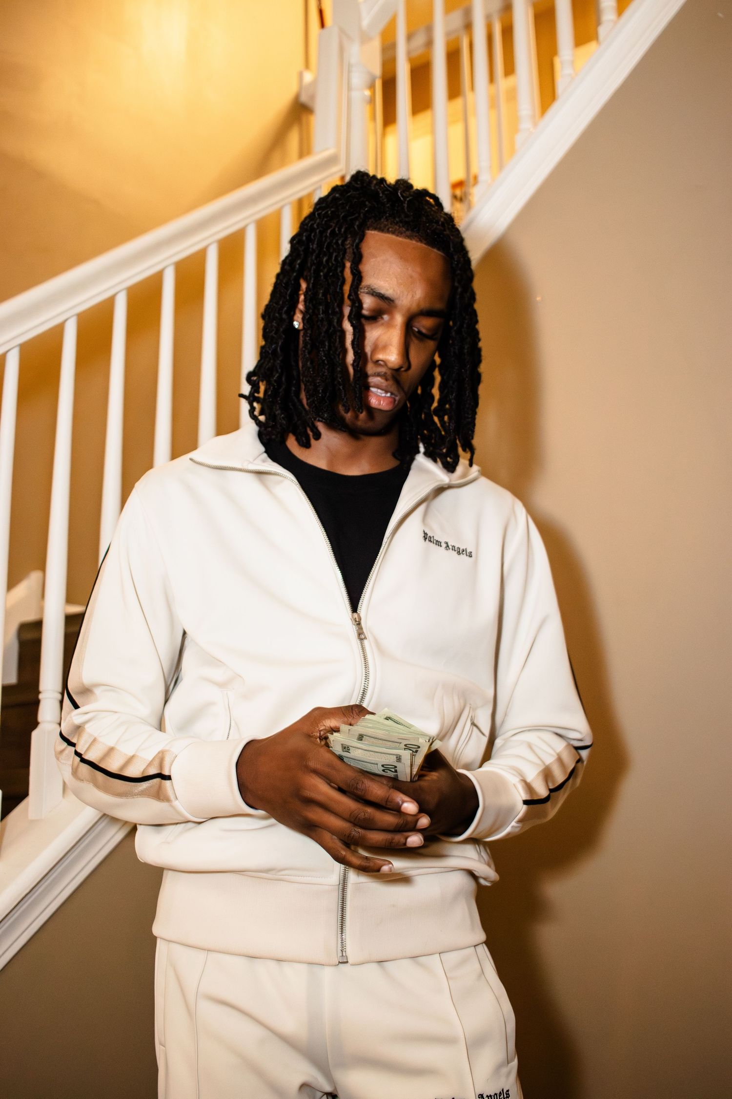 A person in a white outfit looks at their phone while standing against a beige wall with white railings.