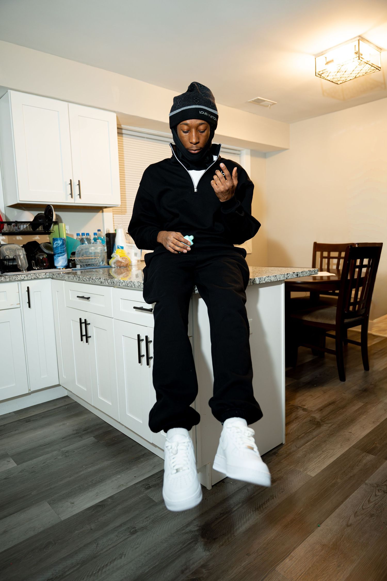 Person in black hoodie and white pants poses casually in modern kitchen with white cabinets and wood flooring.