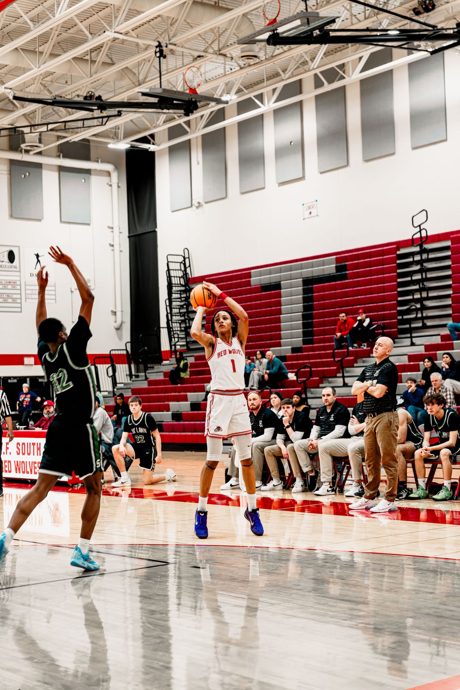 Basketball players practicing jumping drills in a gymnasium with red bleachers and spectators.