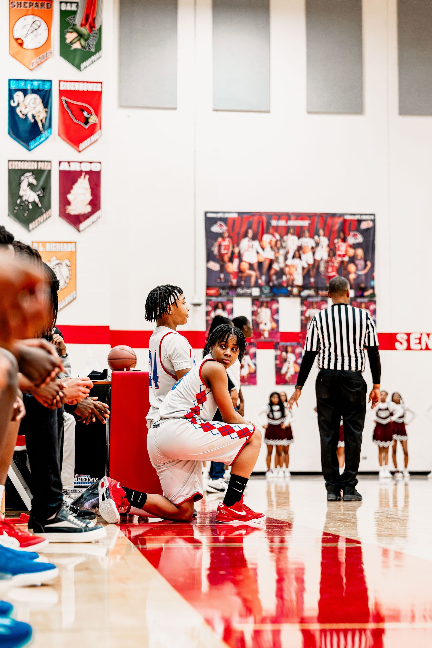 Basketball players sit on the sidelines of a red court during a game break with banners hanging on the wall.
