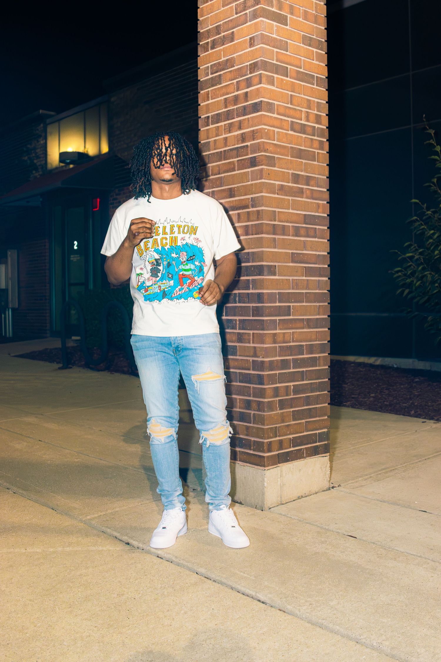 A person wearing ripped jeans and a graphic t-shirt poses against a brick wall at night.