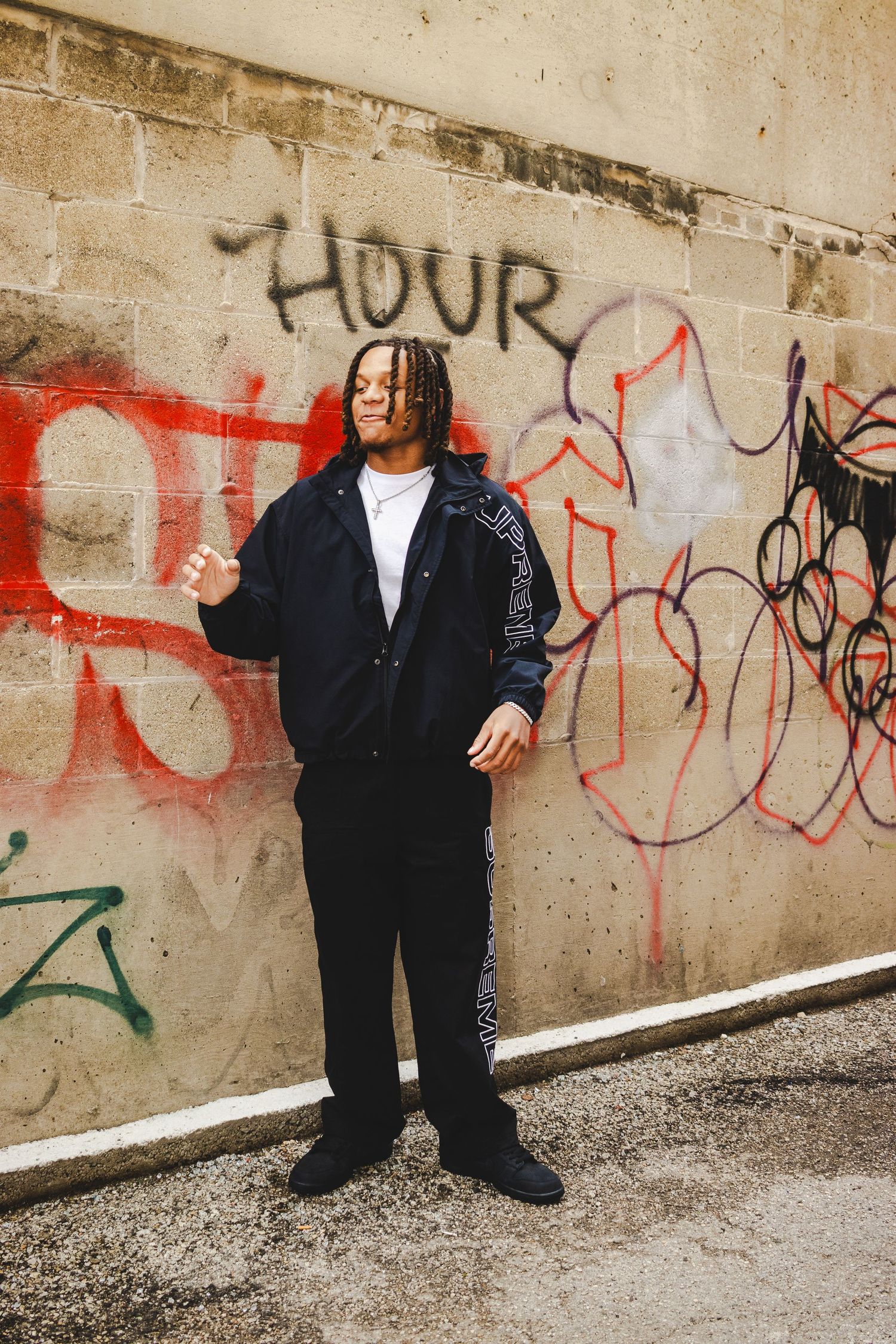 Person in black jacket and pants standing against concrete wall with graffiti art.