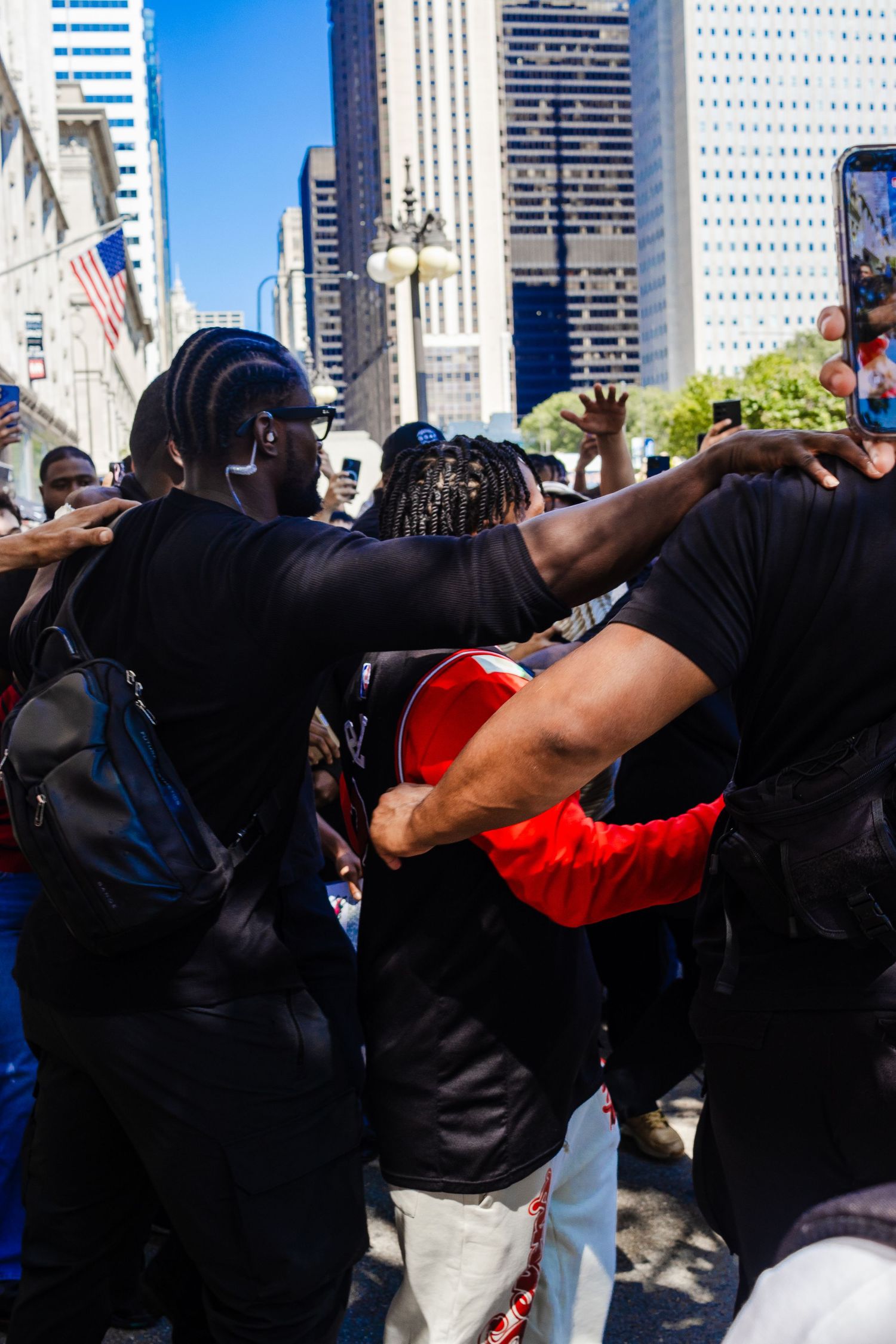 A group of protesters gather in an urban setting with tall skyscrapers visible in the background on a sunny day.