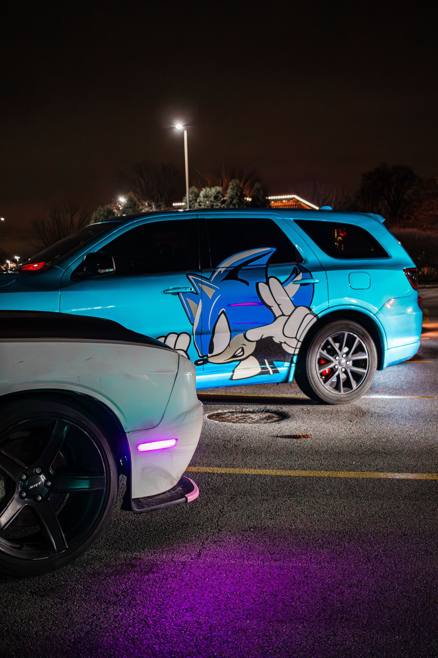 A custom car with Sonic the Hedgehog artwork painted on its side parked at night under street lights with purple ground lighting.