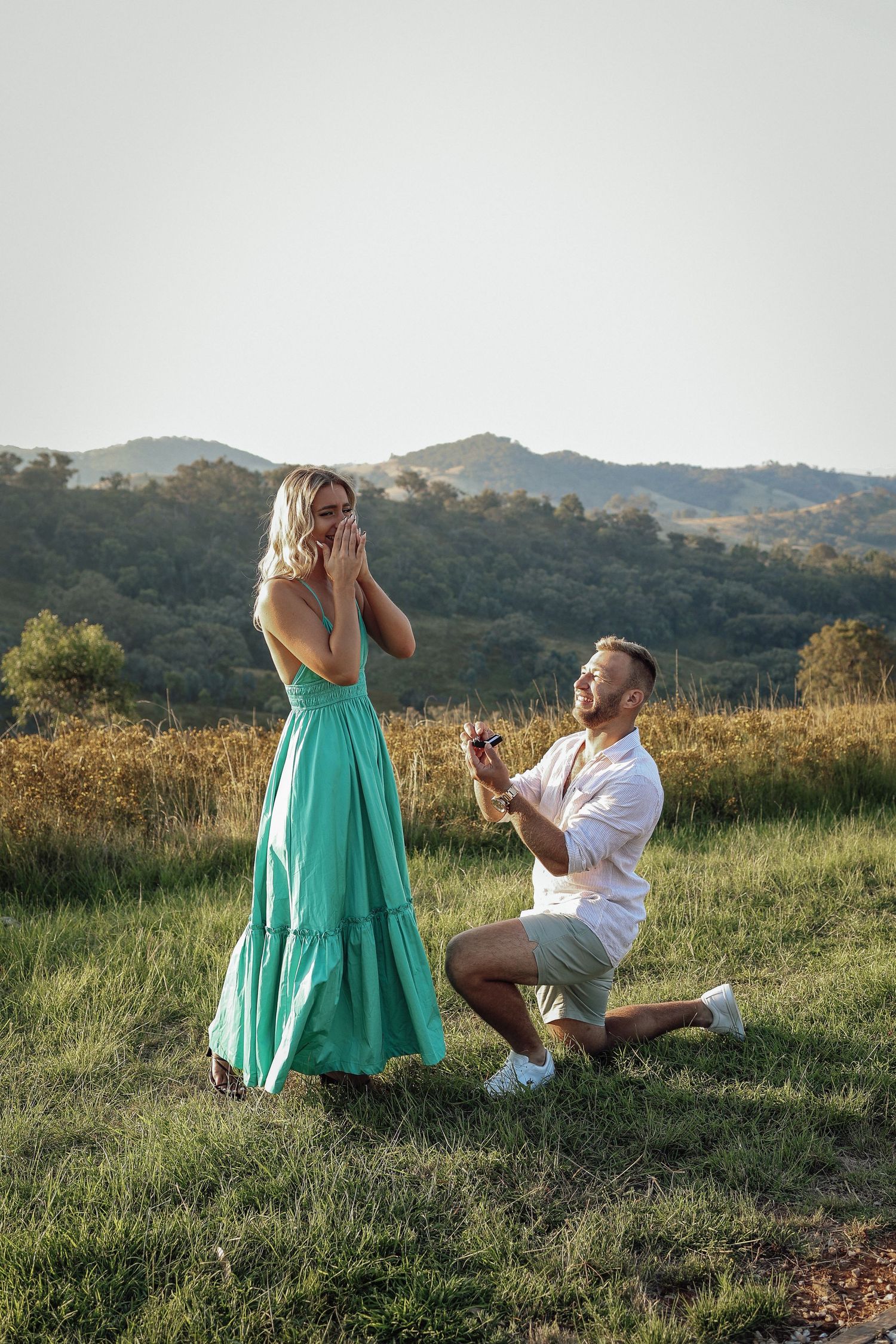 Man proposing on one knee to surprised woman in teal dress in scenic mountain meadow at golden hour.