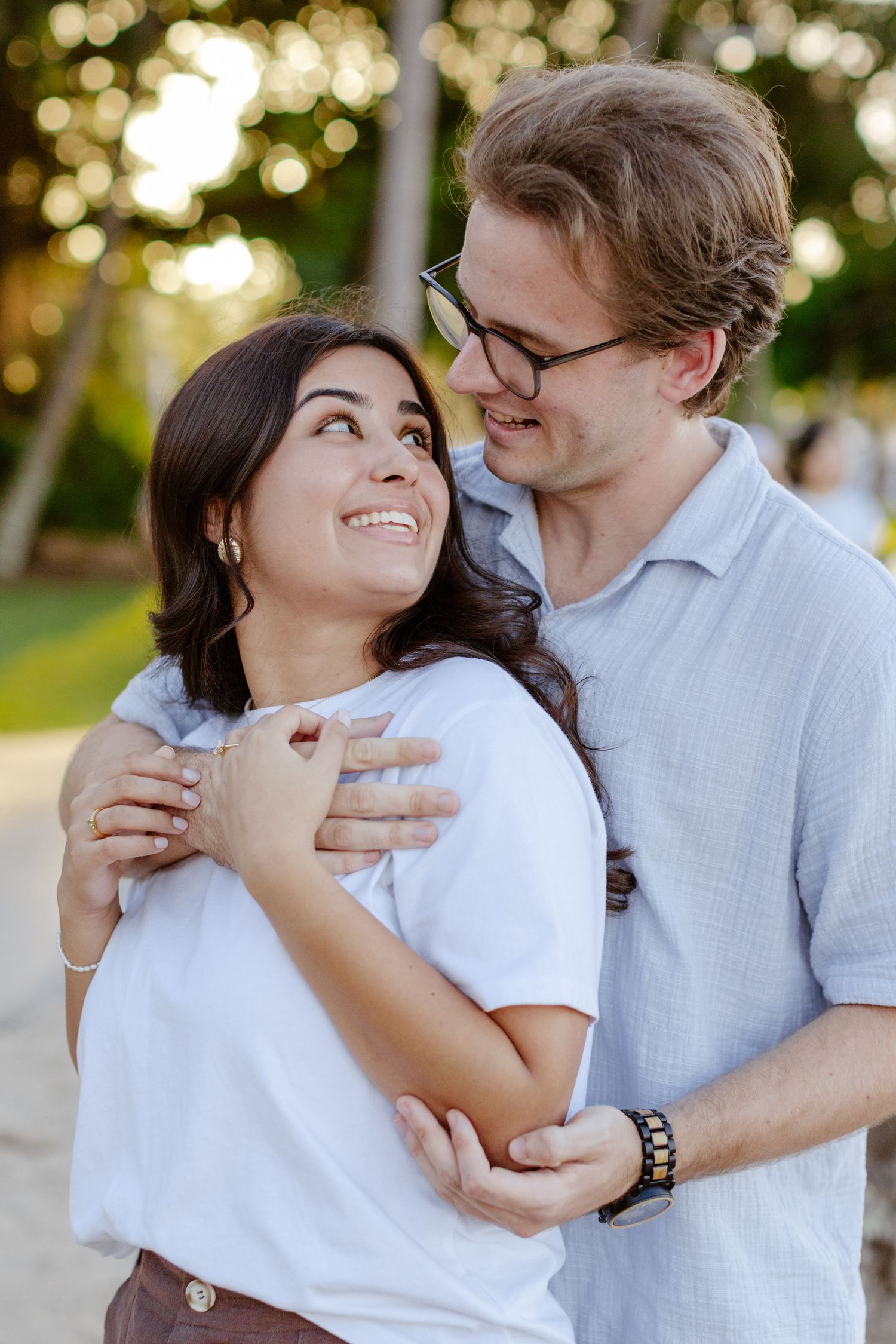 A smiling couple embraces outdoors at sunset, woman in white tee looks up lovingly at man in glasses wearing a blue shirt.
