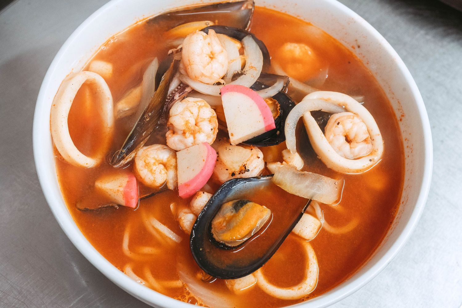 A bowl of seafood soup with shrimp, mussels and squid rings in a vibrant orange-red broth.