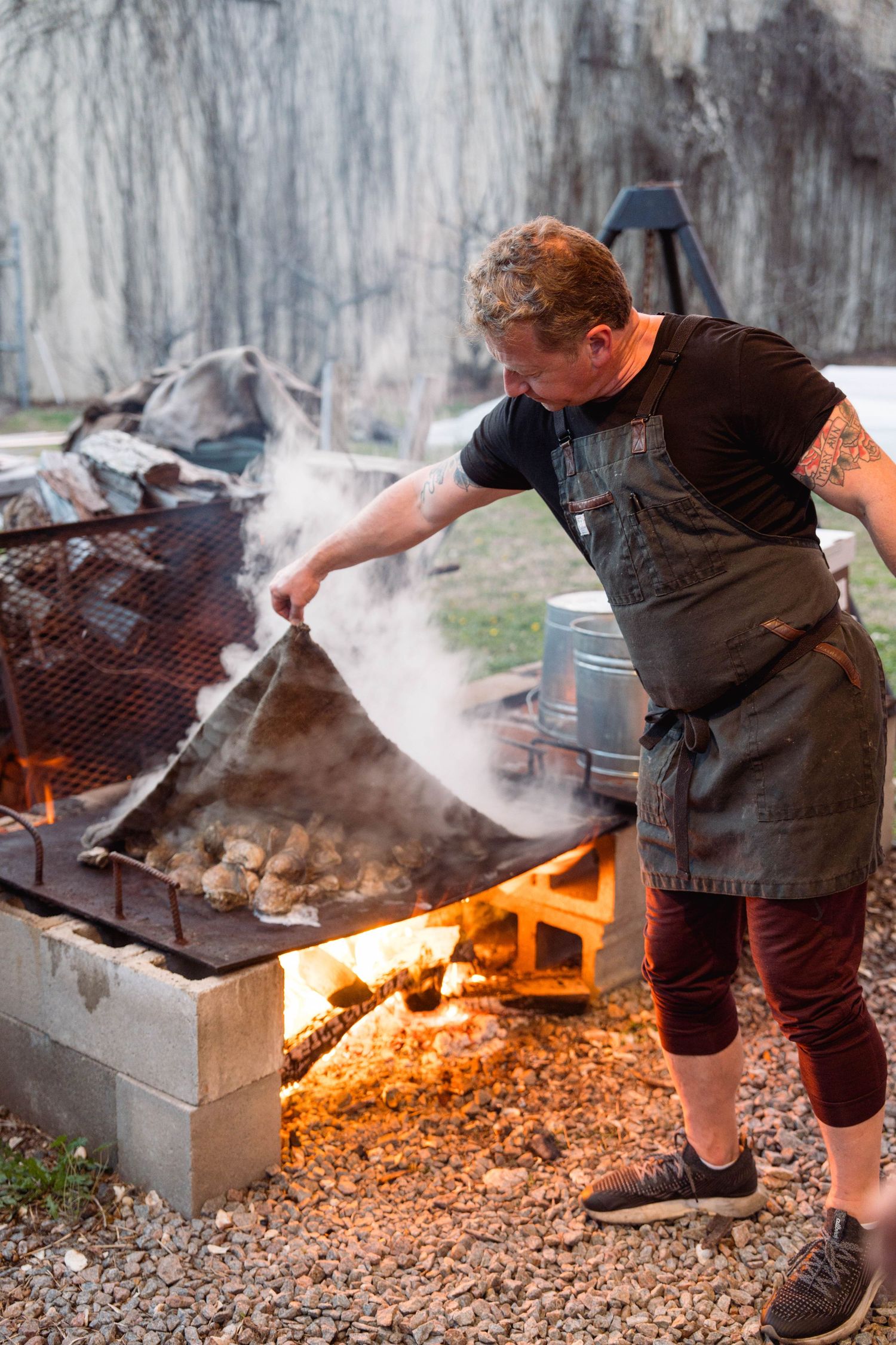 Person grilling meat on an outdoor barbecue grill with glowing embers and rising smoke.