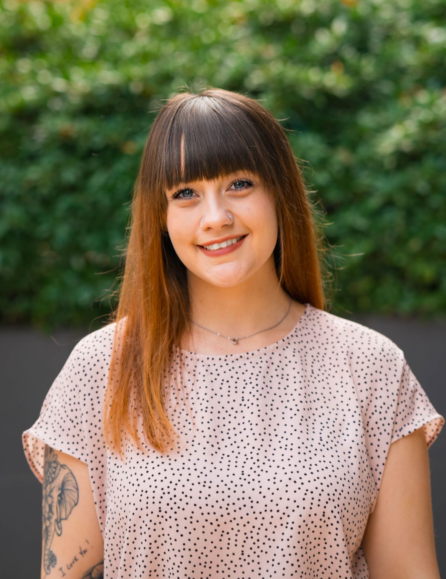 Woman wearing polka dot blouse smiles while sitting outdoors against green foliage background.