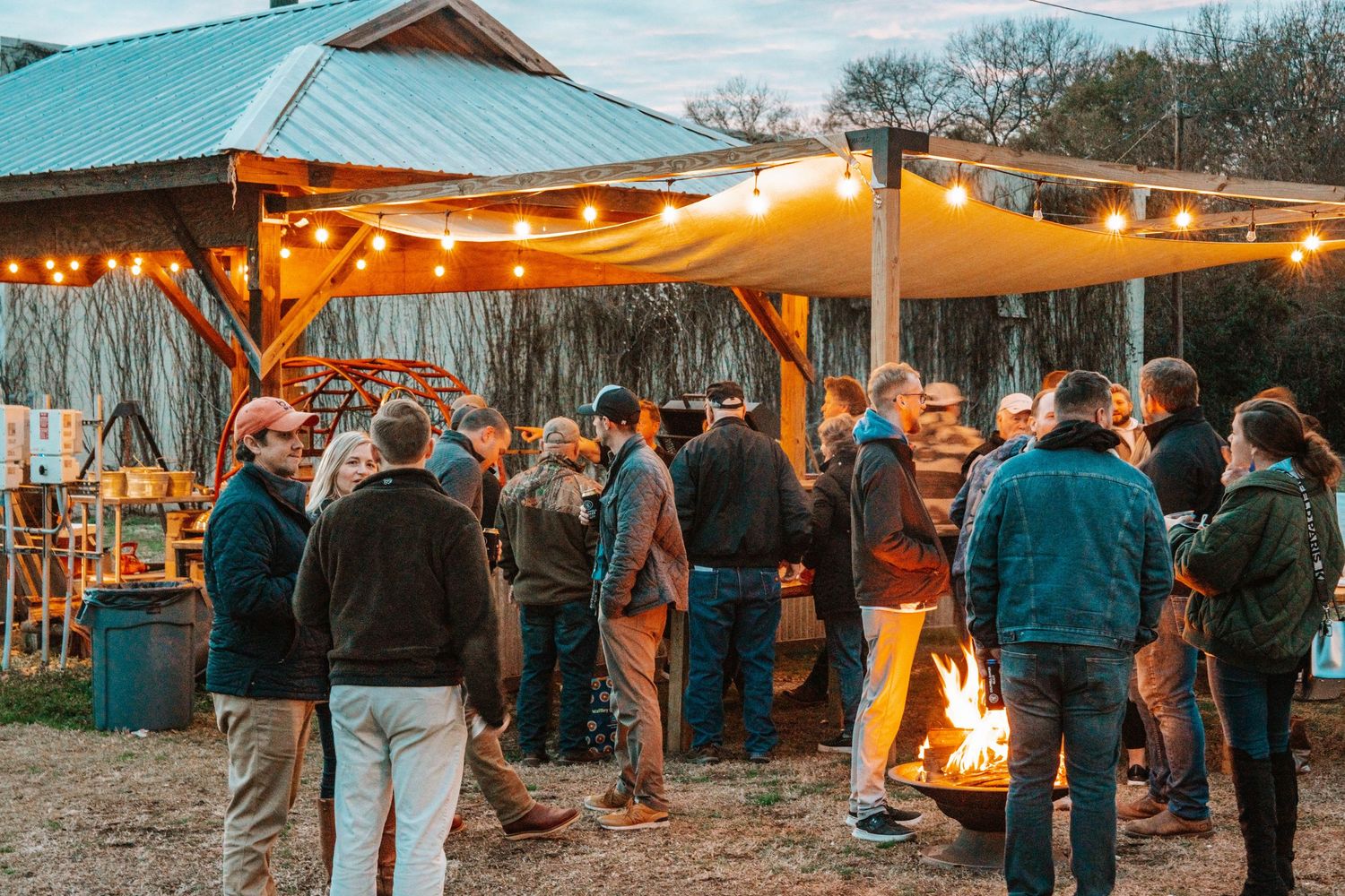People gather under string lights at an outdoor evening event with a fire pit.