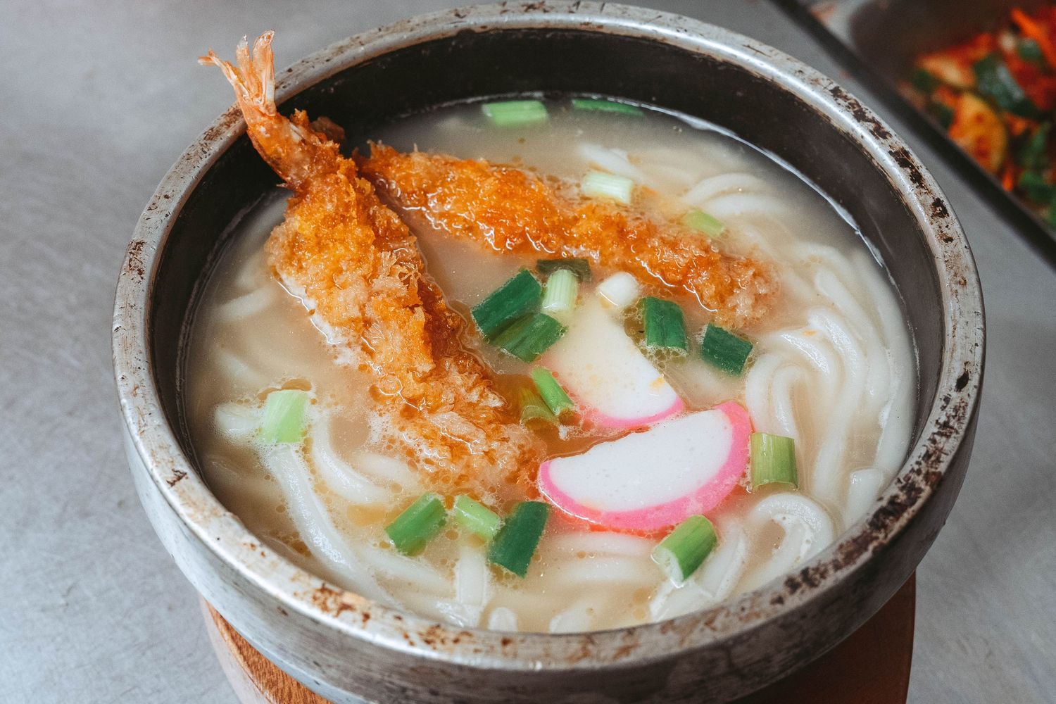 A bowl of udon noodle soup garnished with tempura shrimp, green onions, and sliced radish in a creamy broth.