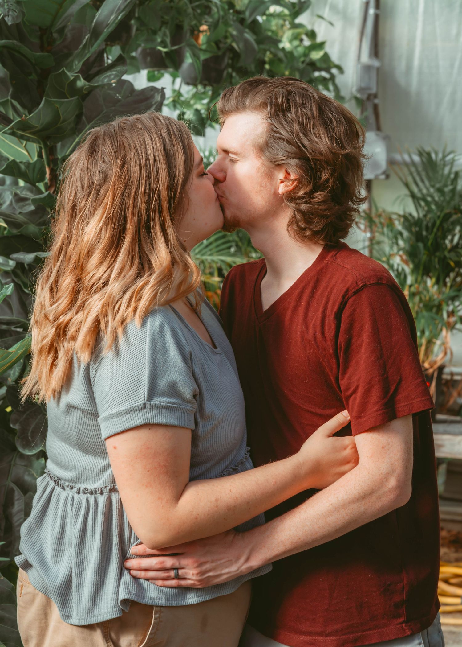 Romantic couple in casual attire embracing in an indoor setting with lush houseplants.