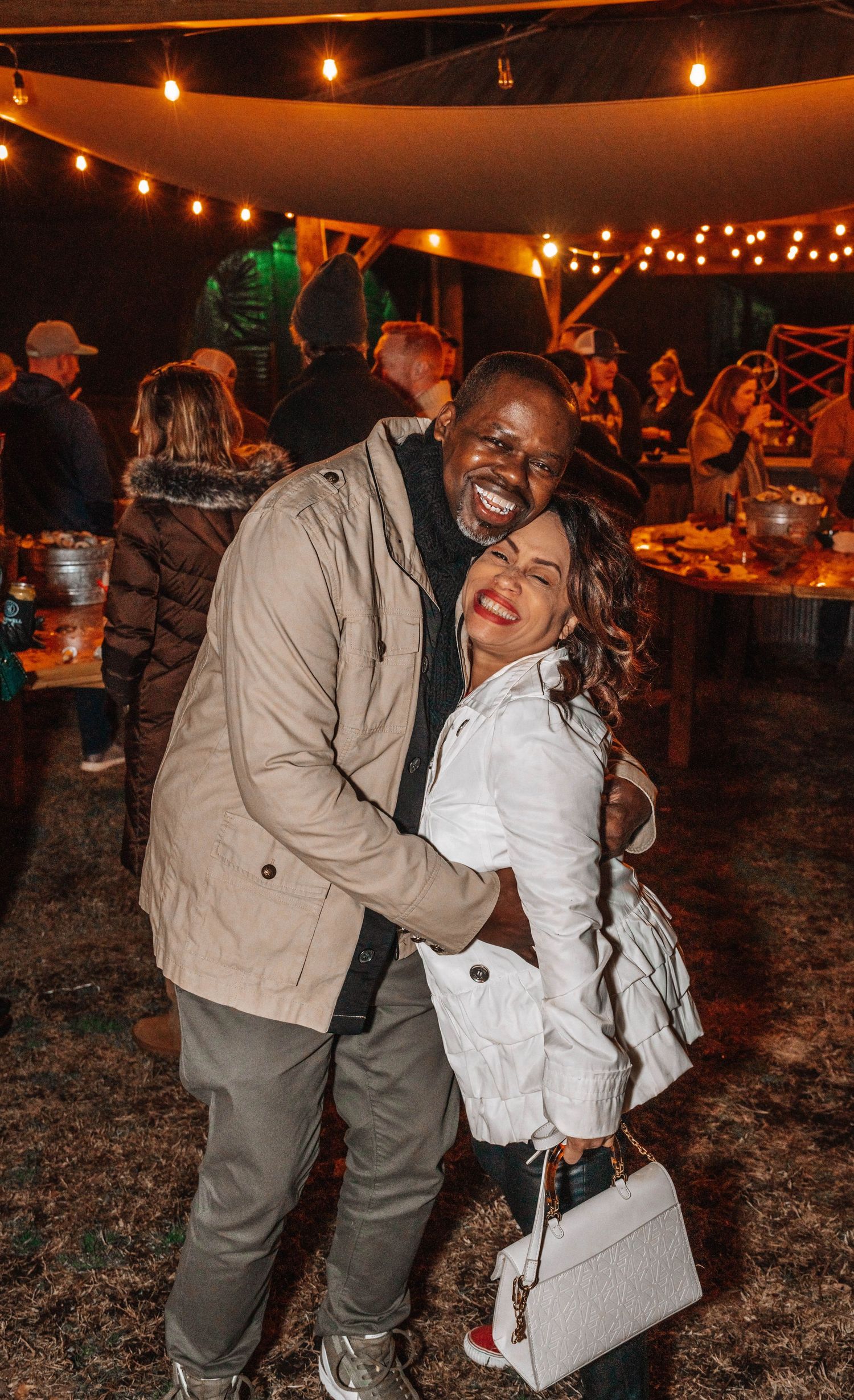 A happy couple embraces and laughs at an outdoor evening event under string lights.