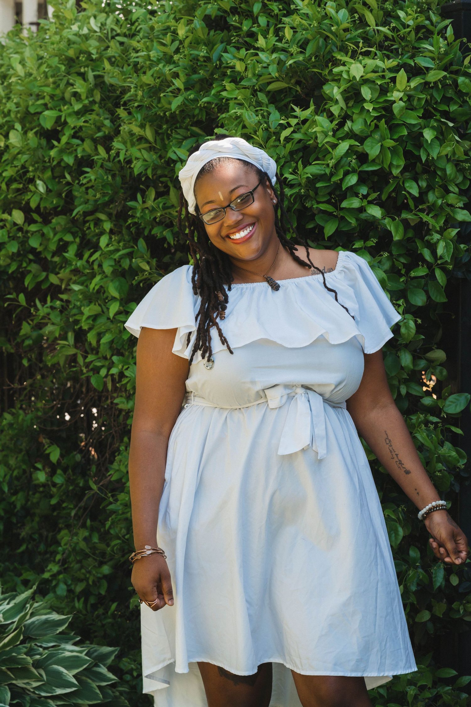 A white ruffled summer dress with scalloped details shown in multiple angles against a green hedge background.