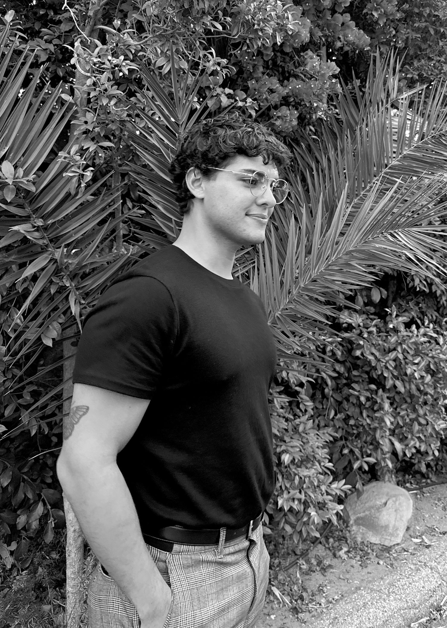 A person in a black t-shirt poses against a tropical garden backdrop in a black and white photo series.