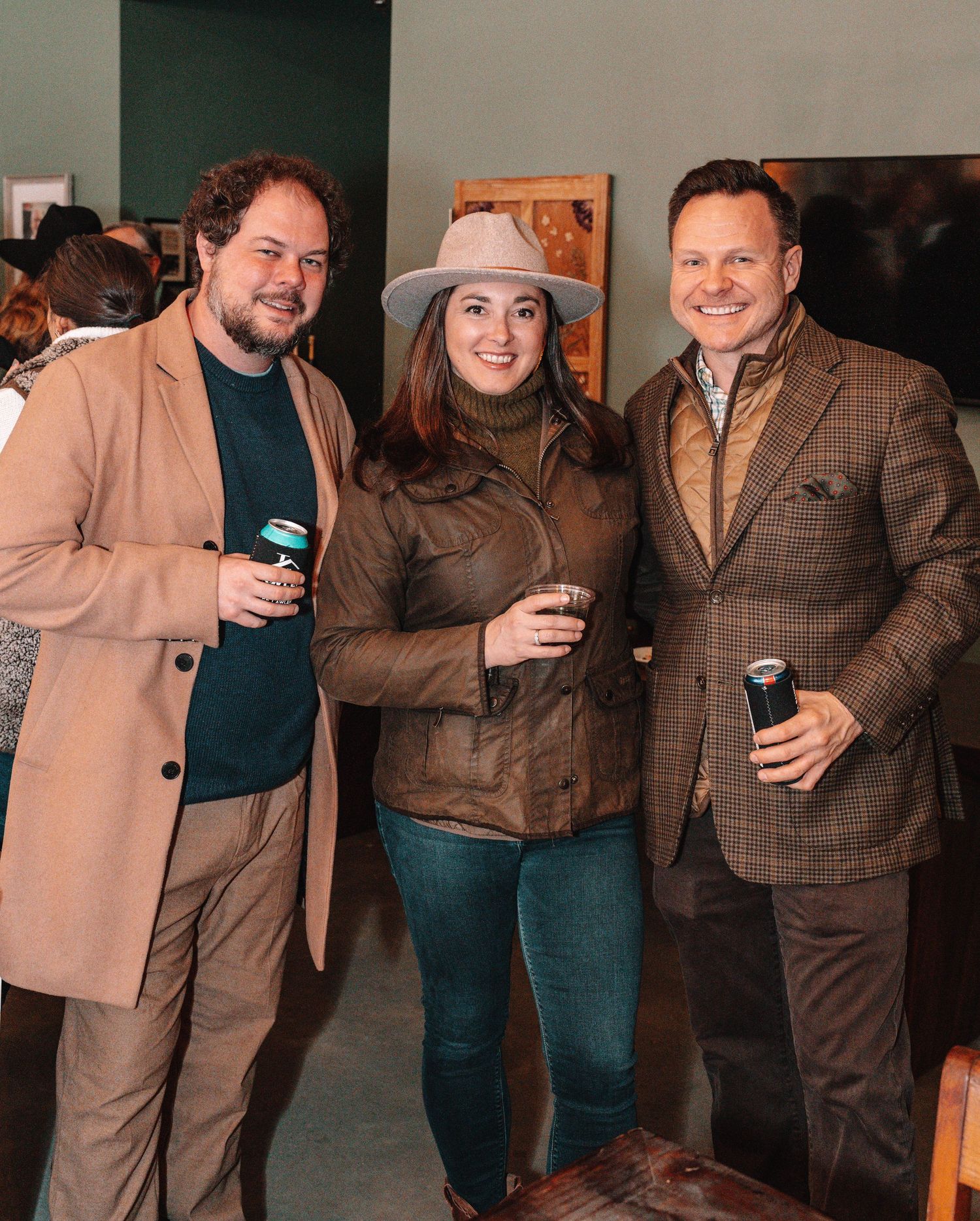 Three friends pose together at an indoor social gathering holding beverages.