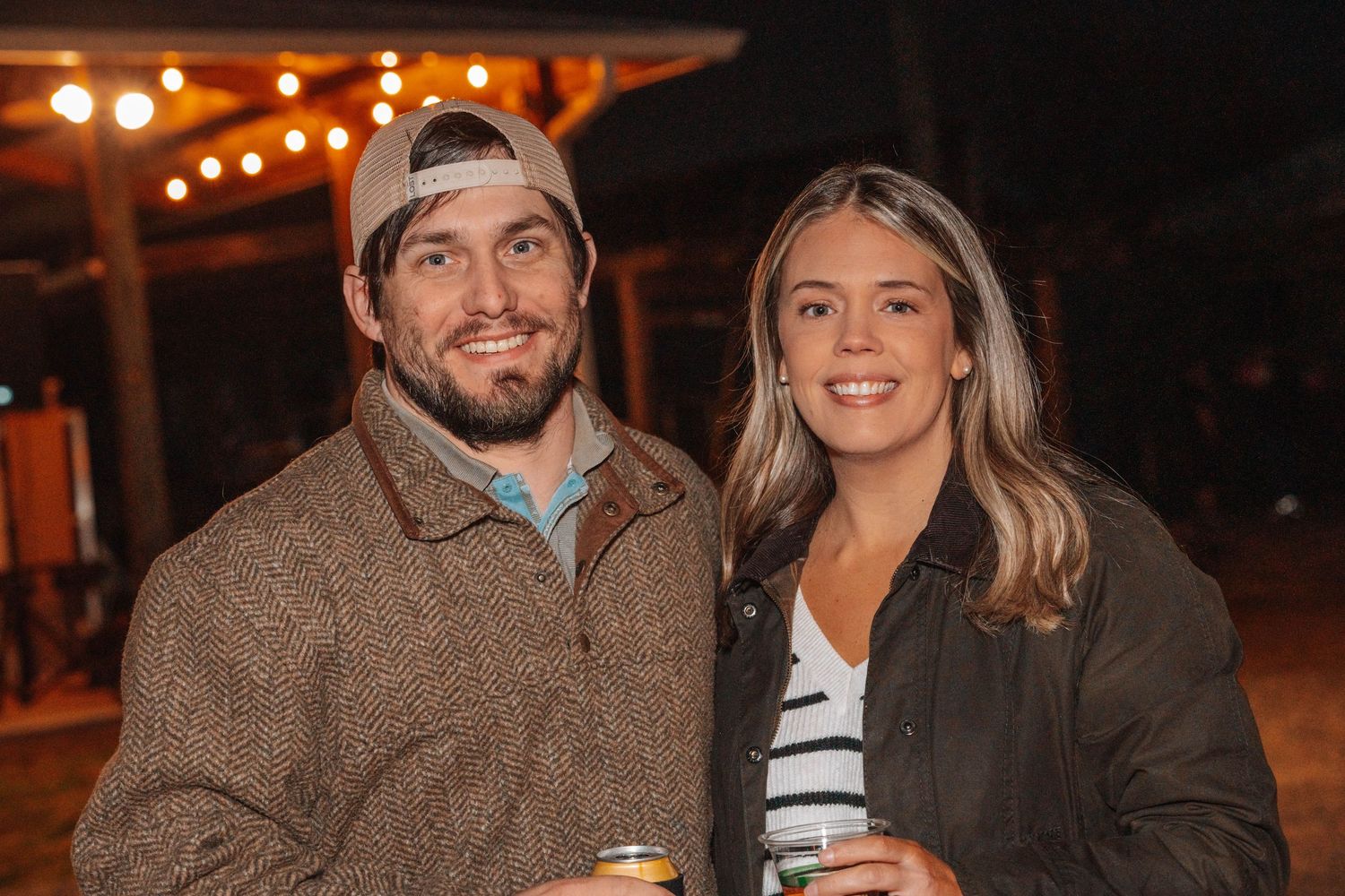 A couple smiles at the camera under outdoor string lights at night.